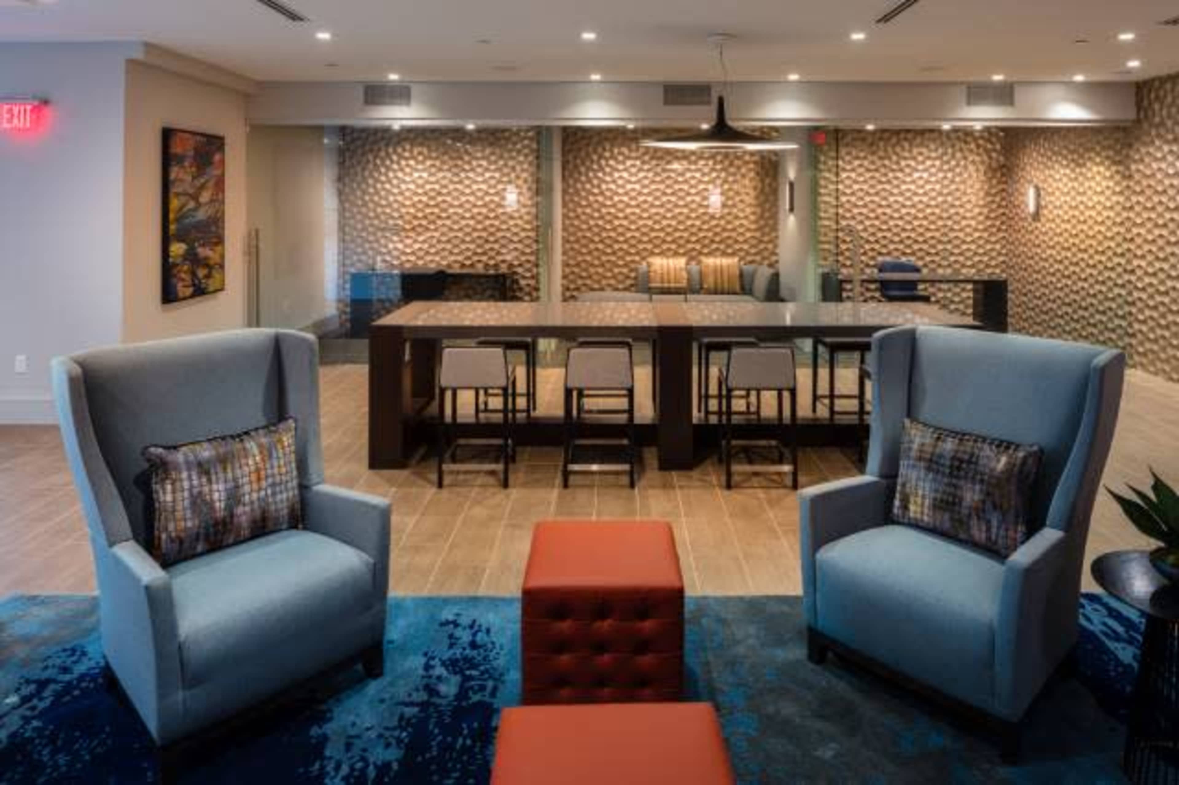 The image shows a modern lounge area with two blue upholstered chairs, a red ottoman, and a long communal table surrounded by bar stools against a textured wall.