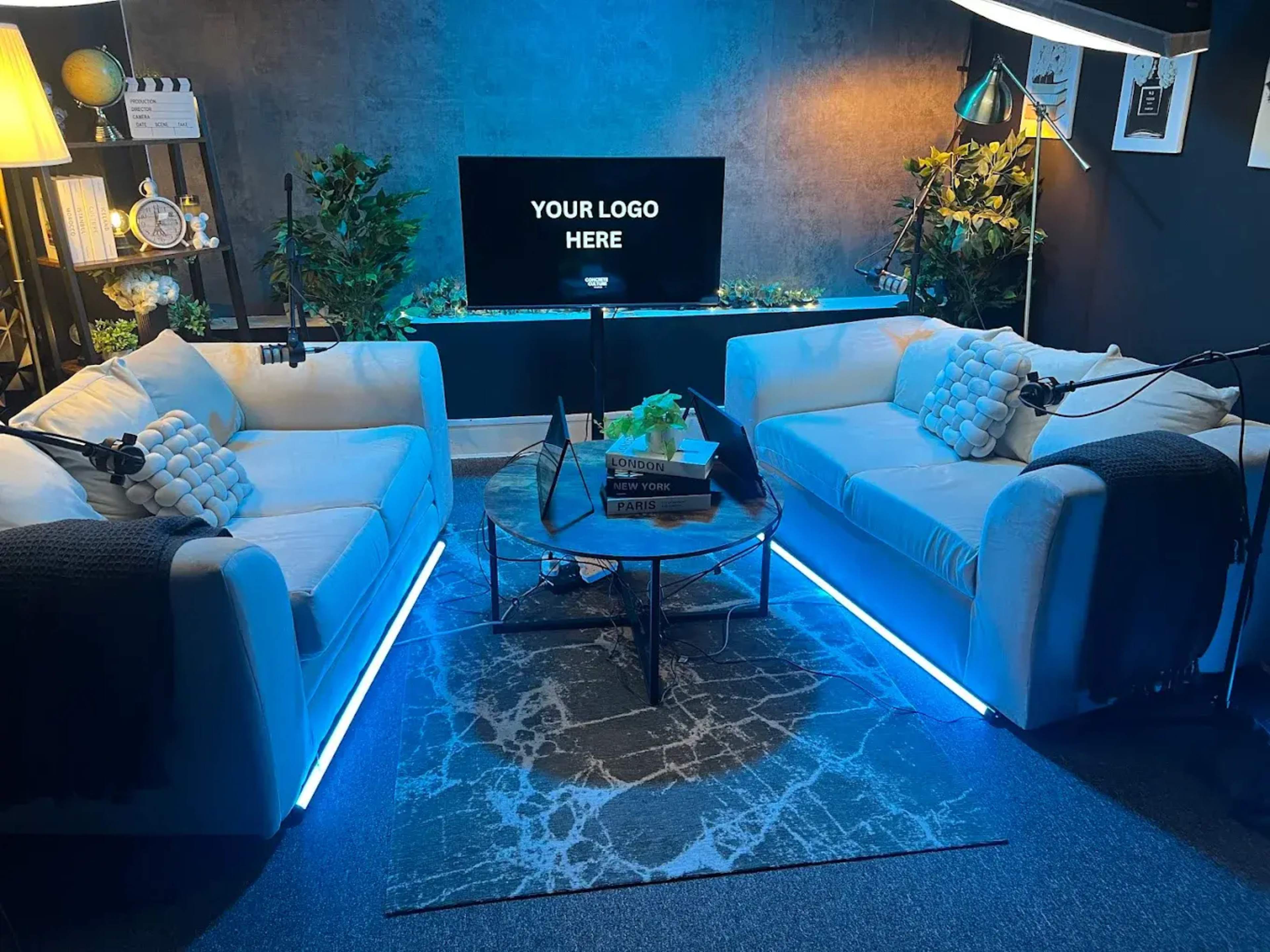 The image shows a modern lounge setup featuring two white sofas, a coffee table with decorative items, and a television displaying a placeholder for a logo, all illuminated by ambient lighting.