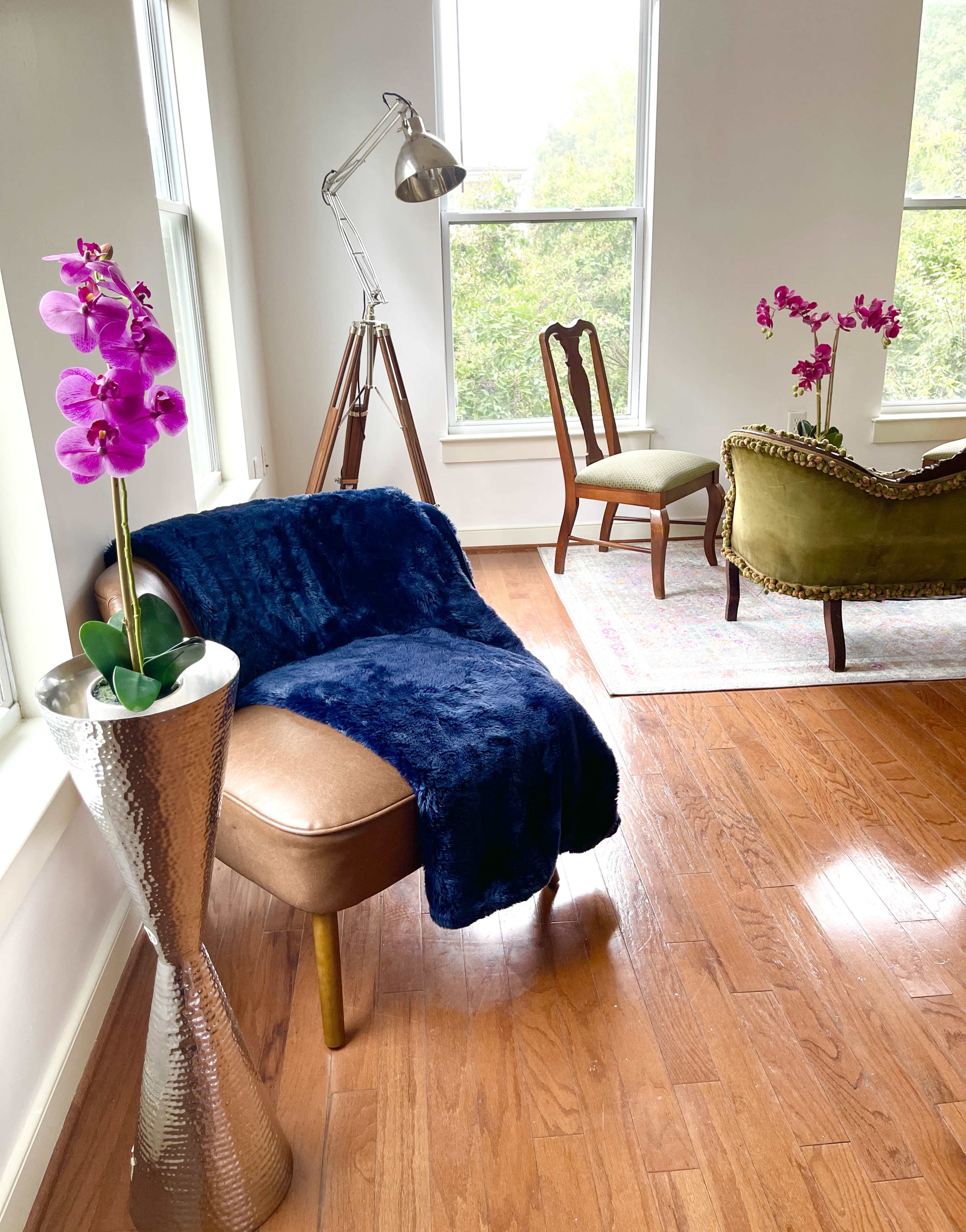 The image shows a cozy living space featuring a blue upholstered chair with a blanket, a tall metallic vase with a pink orchid, a tripod lamp, and a green upholstered chair with a wooden frame.