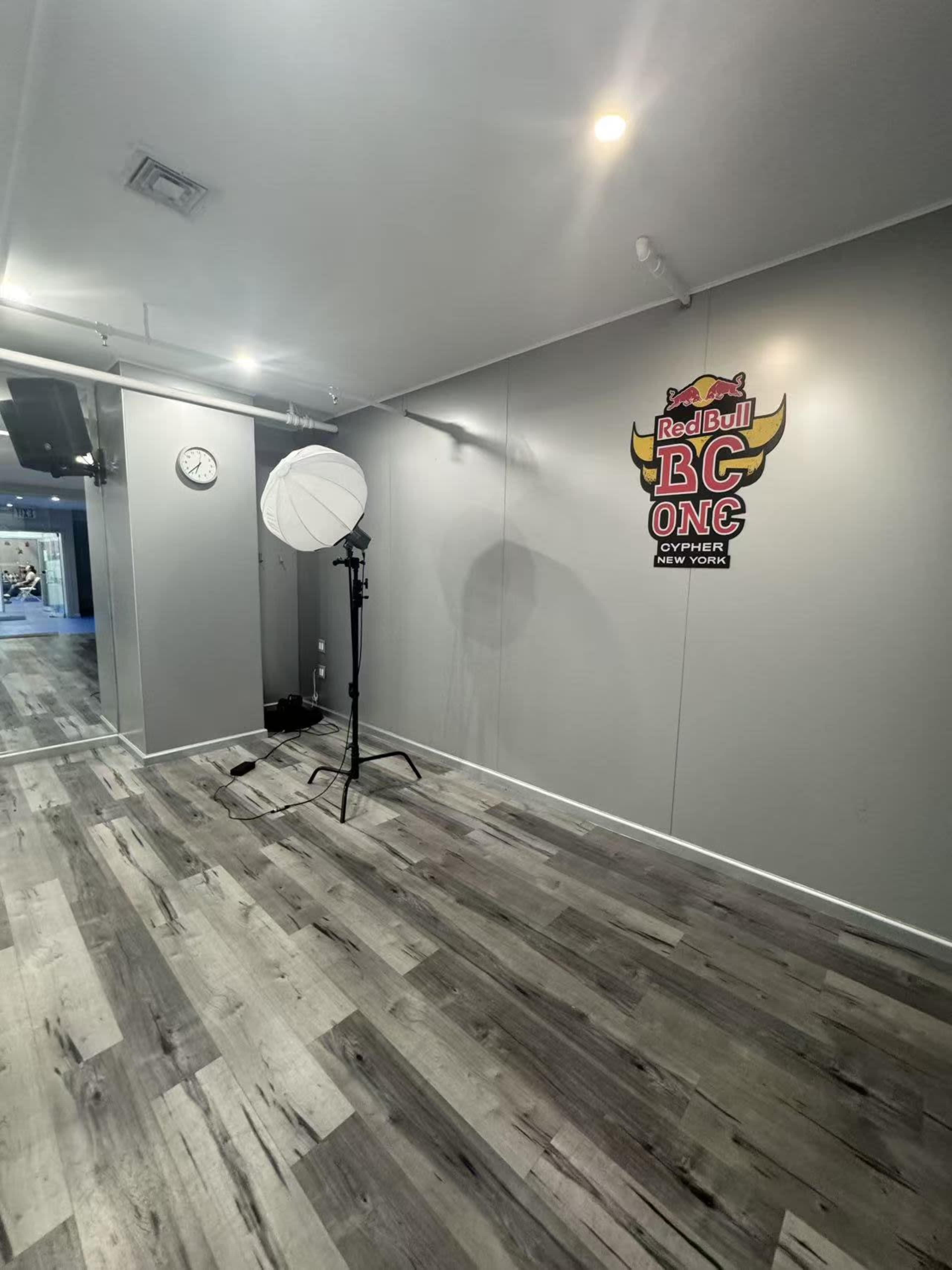 The image shows a sparsely furnished room with gray walls, wooden flooring, a clock on the wall, and a light stand with a softbox.