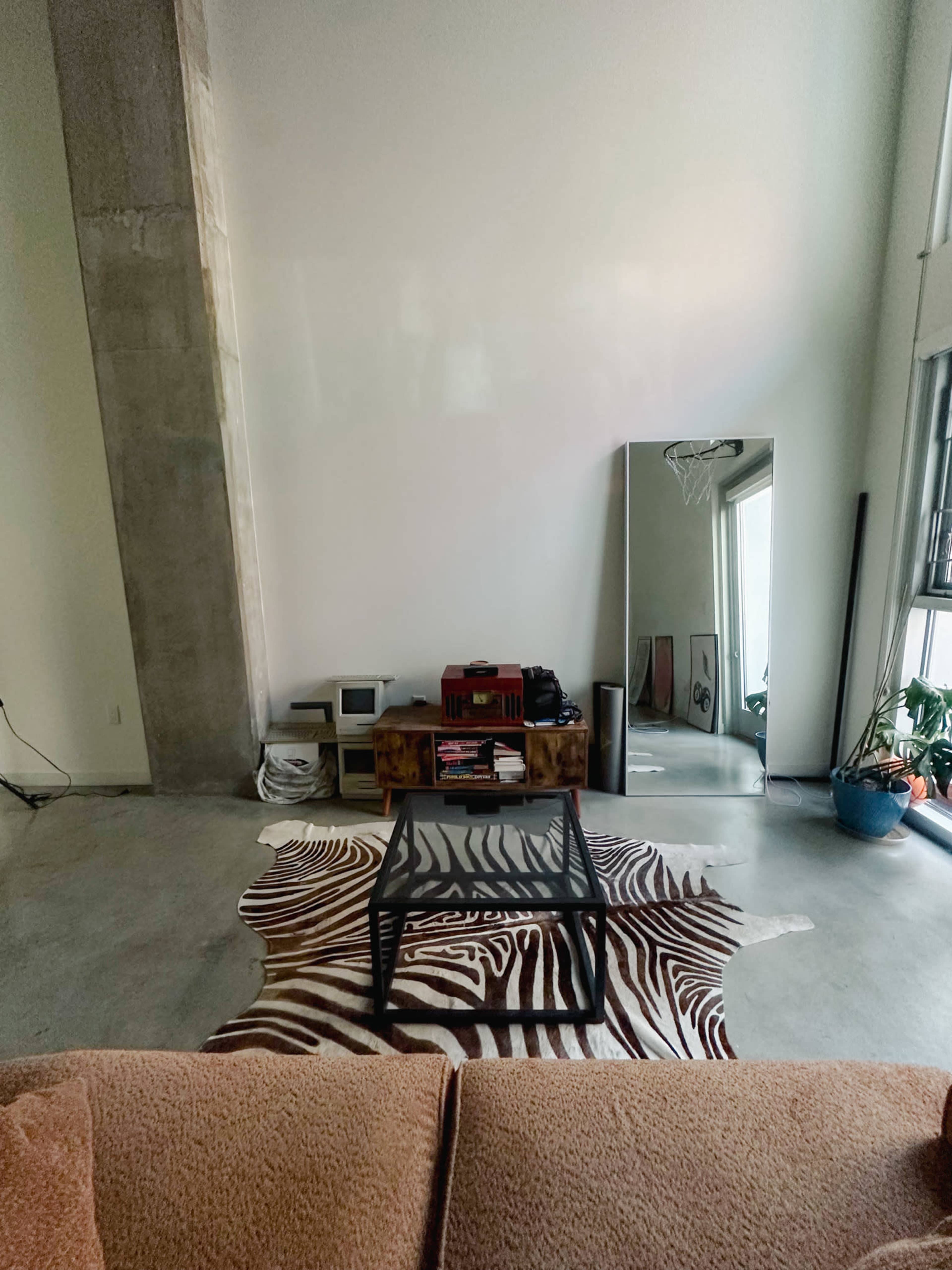 The image shows a minimalist living room with a large mirror, a glass coffee table on a zebra-striped rug, and various furniture and decorations against a plain wall.