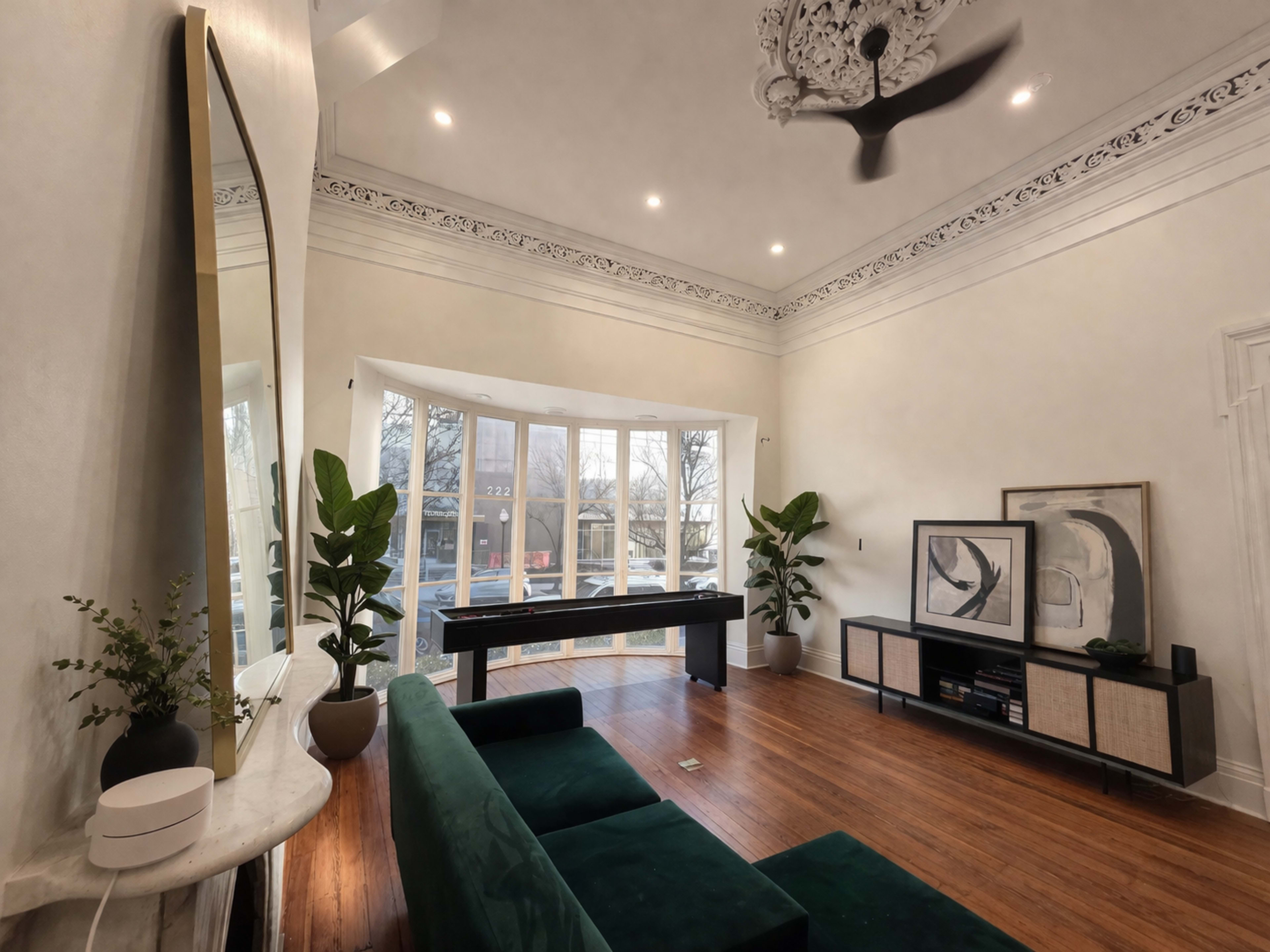 The image shows a spacious living room with a green sofa, a wooden console table, large windows overlooking a balcony, and decorative plants.