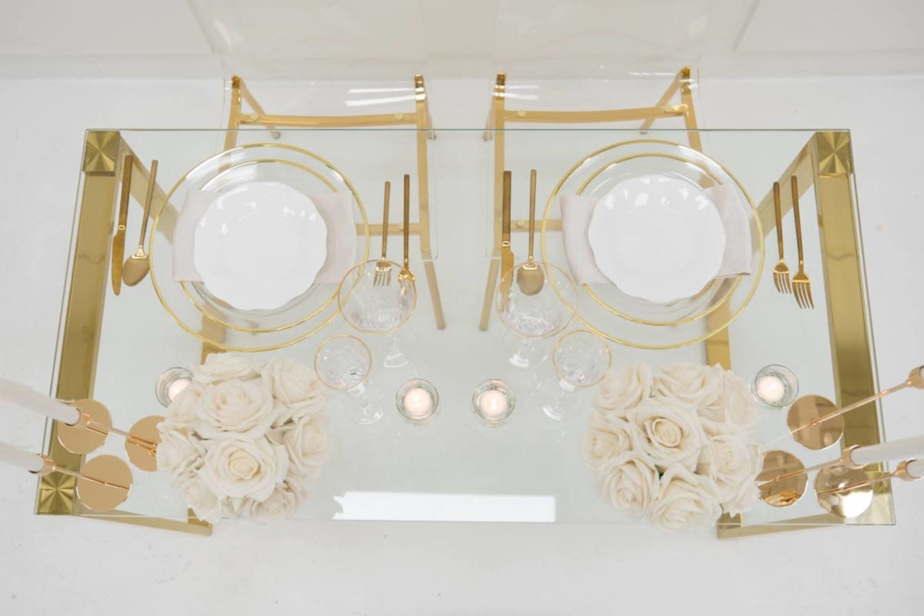 The image shows a glass dining table set with two place settings, gold utensils, clear crystal glasses, and white roses in elegant arrangements.