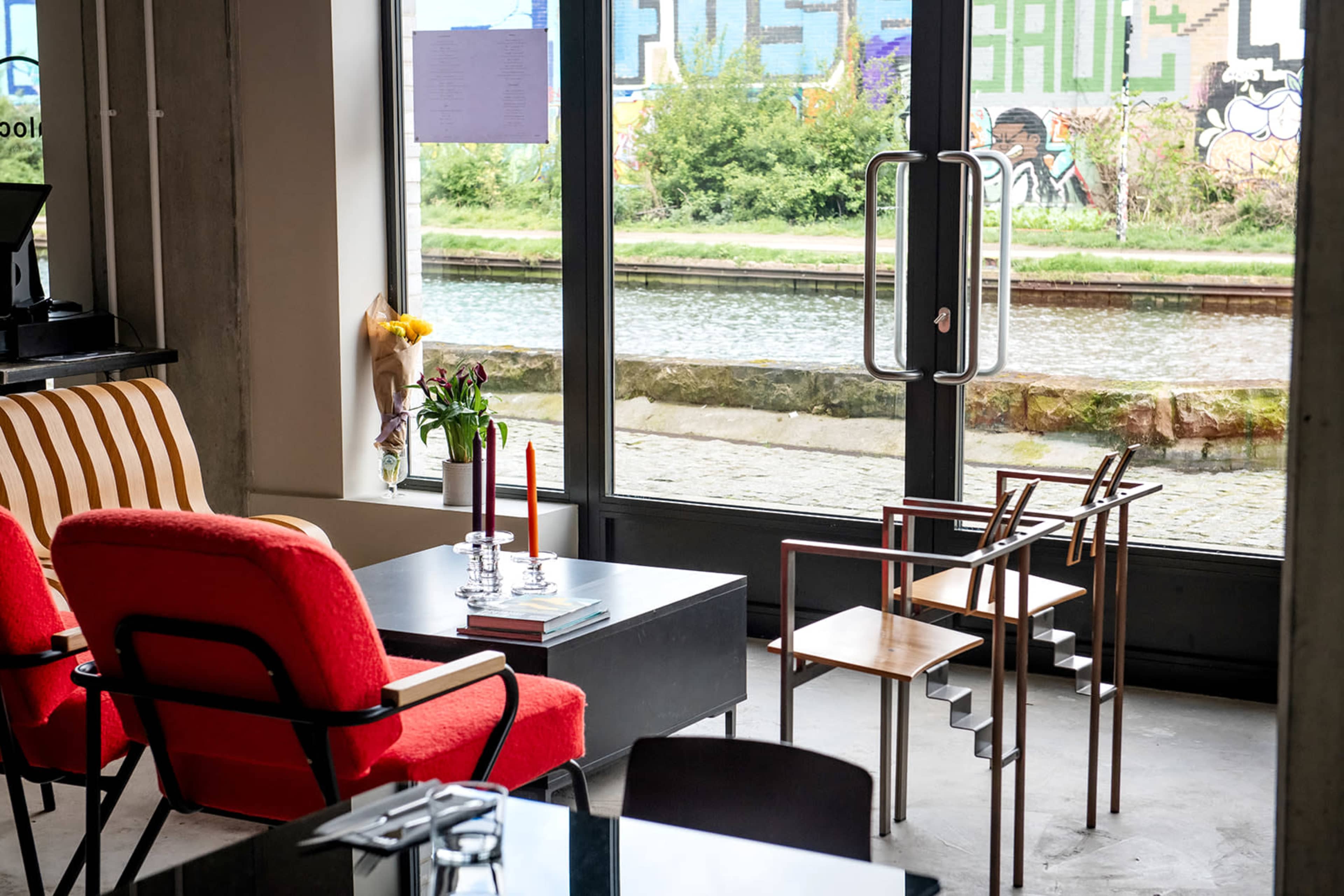 A modern seating area features red and wooden chairs beside a glass door overlooking a canal with graffiti on the opposite bank.