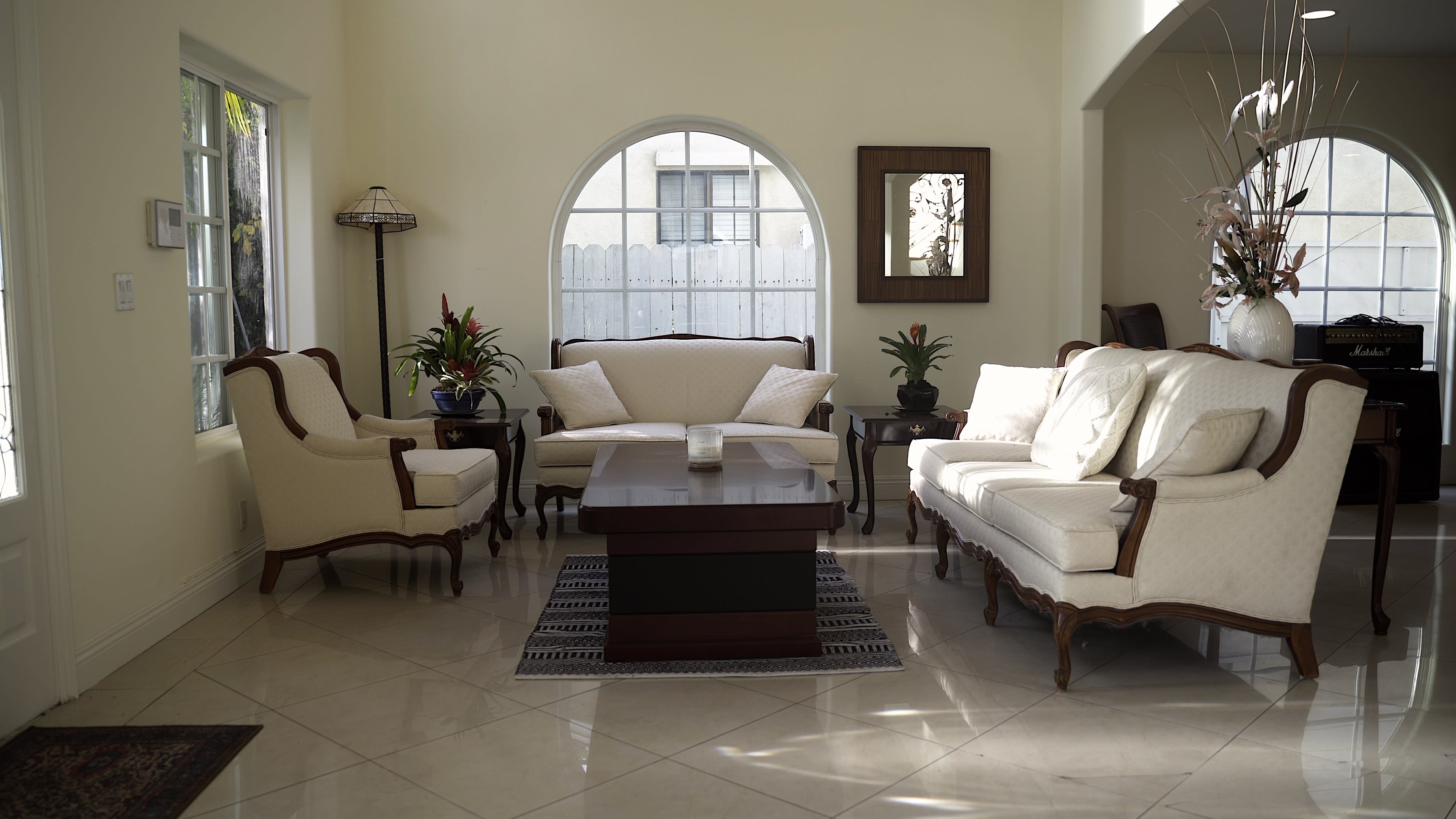 A living room with a set of light-colored seating arranged around a dark coffee table, with potted plants in the corners and a large window letting in natural light.