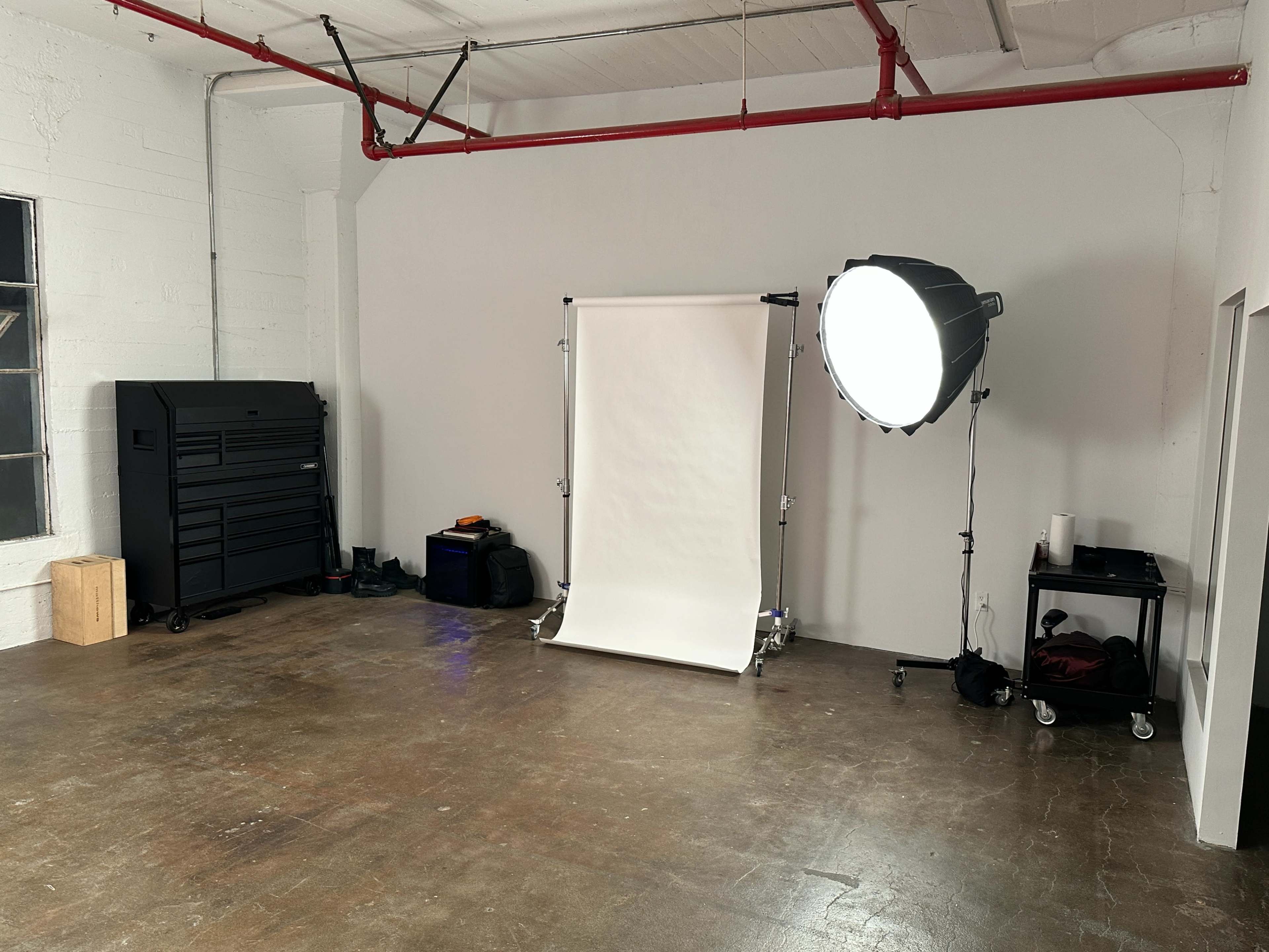 The image shows a spacious room with a wooden floor, featuring a backdrop stand with a white sheet, a large studio light, and a storage unit along the wall.