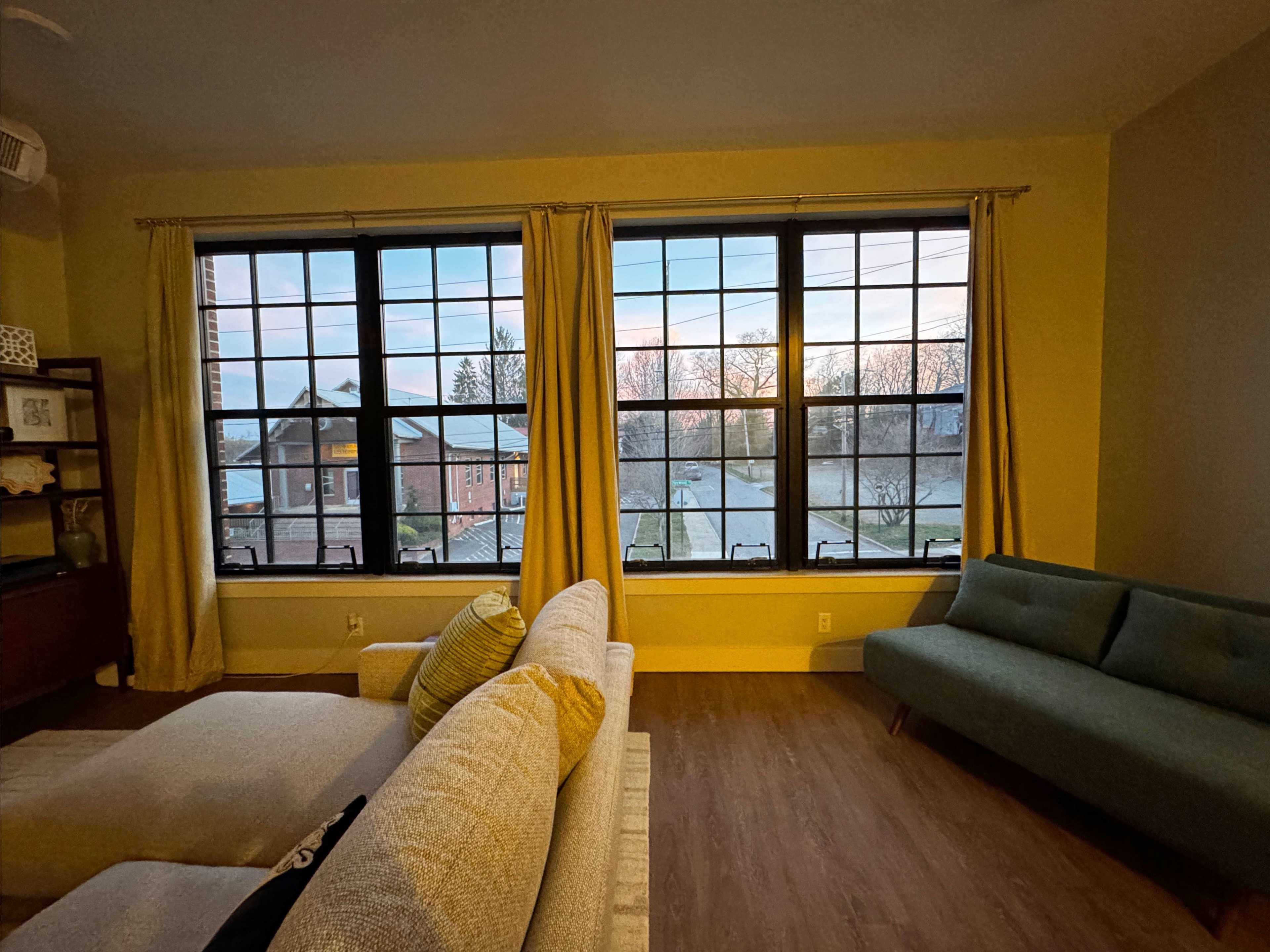 The image shows a cozy living room with large windows revealing a street view at dusk, featuring a sofa and a decorative bookshelf.