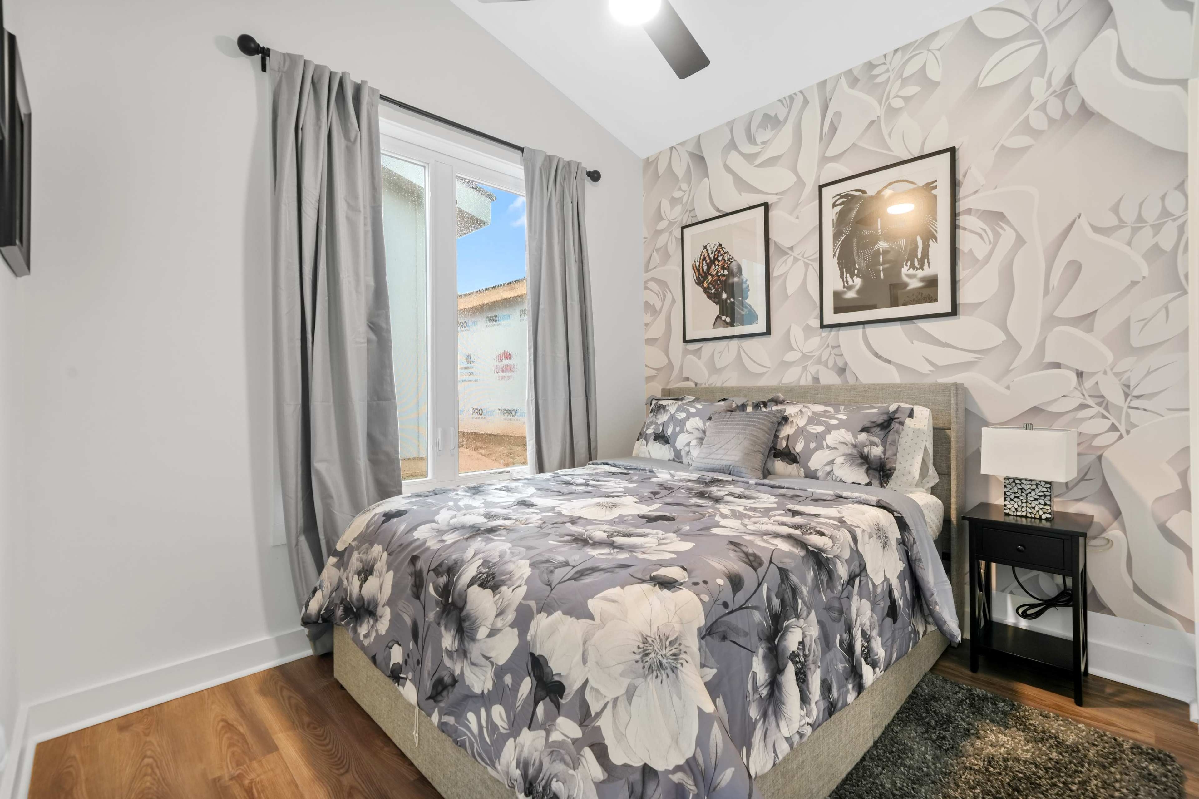 A bedroom features a bed with floral-patterned bedding, two framed pictures on the wall, and a window with curtains.