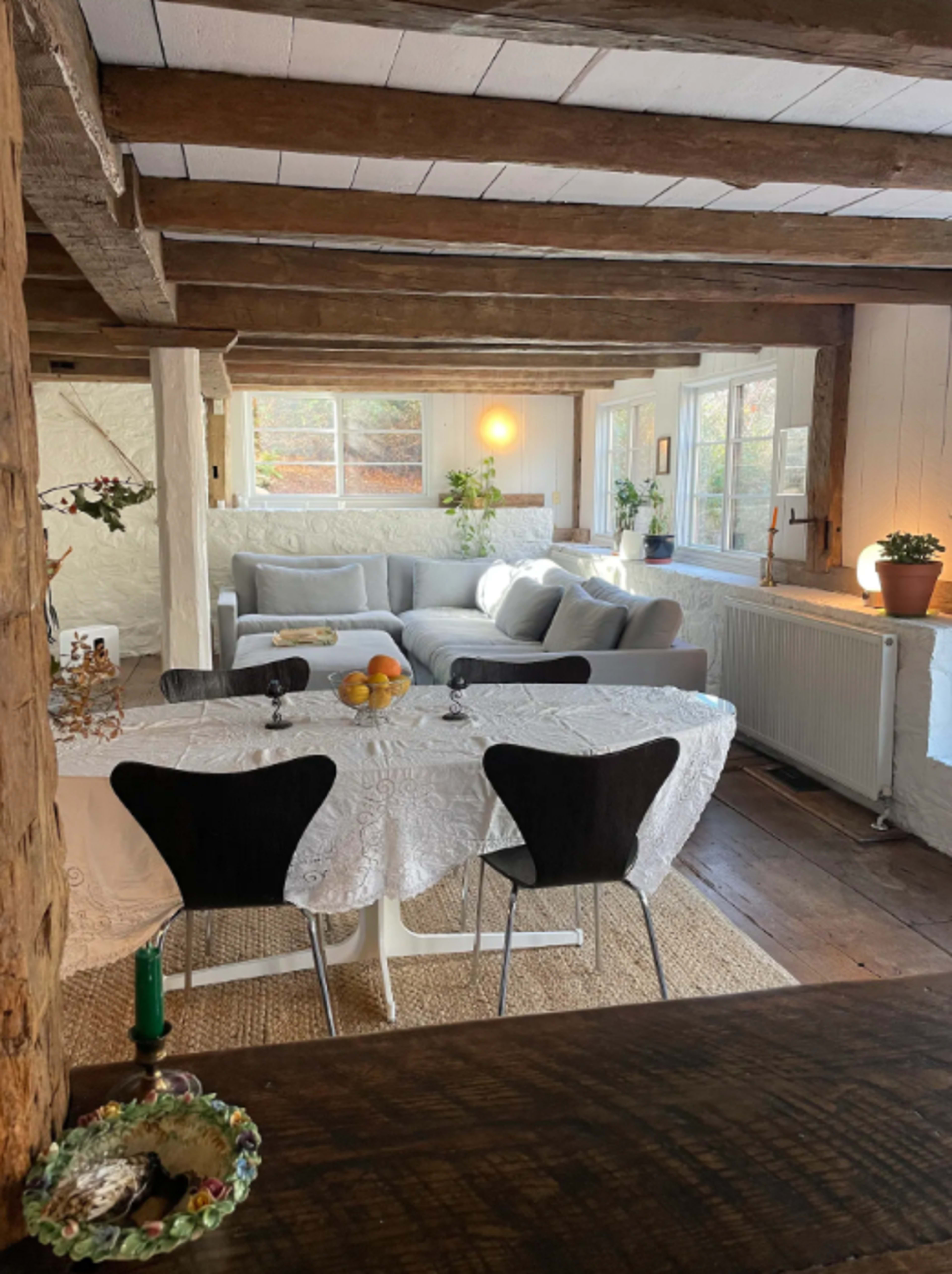 A bright room features a dining table with a white tablecloth, black chairs, and a living area with a gray sofa and large windows.