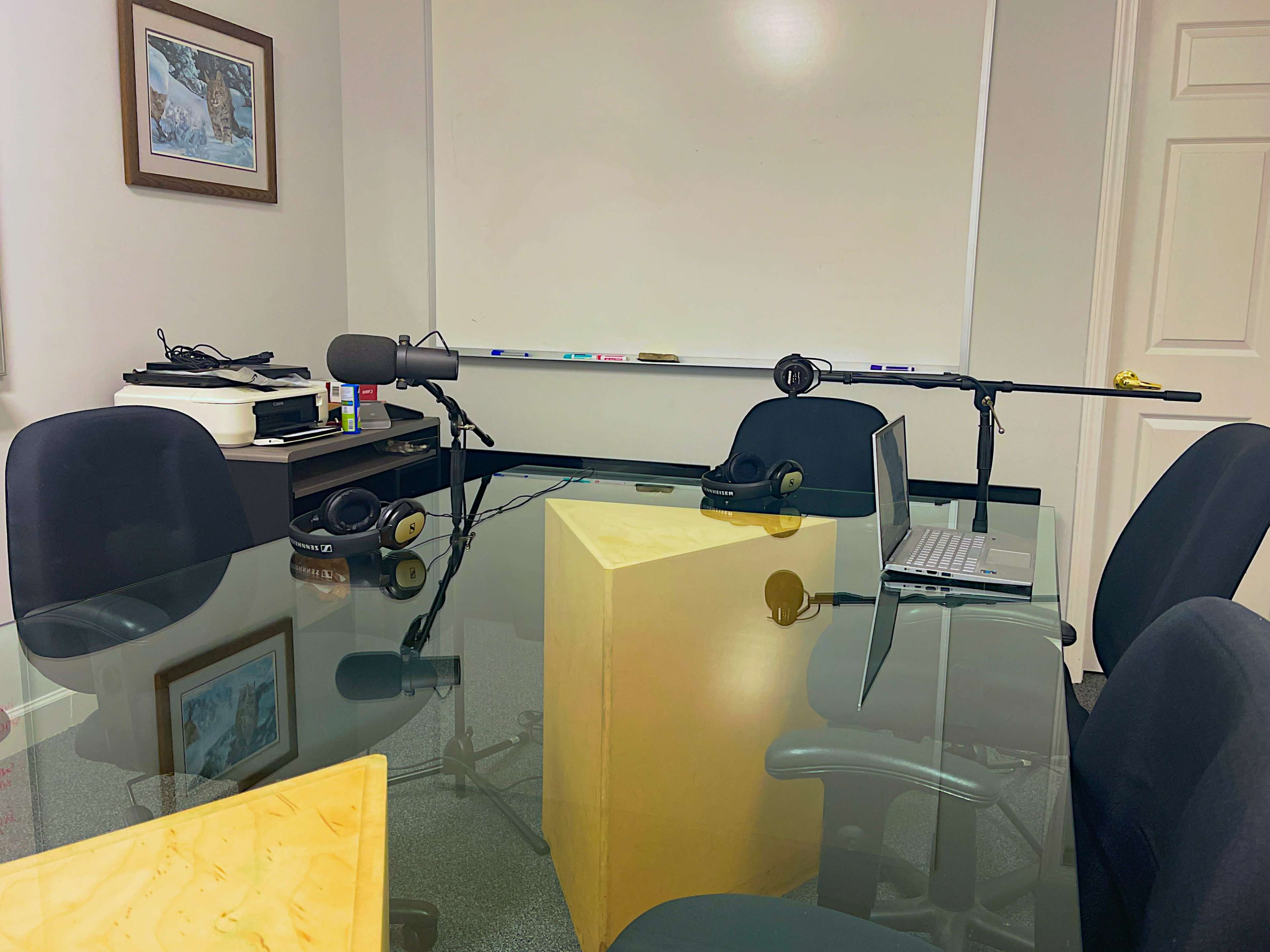 A conference room features a glass table, black chairs, a whiteboard, and equipment for recording, including microphones and headphones.