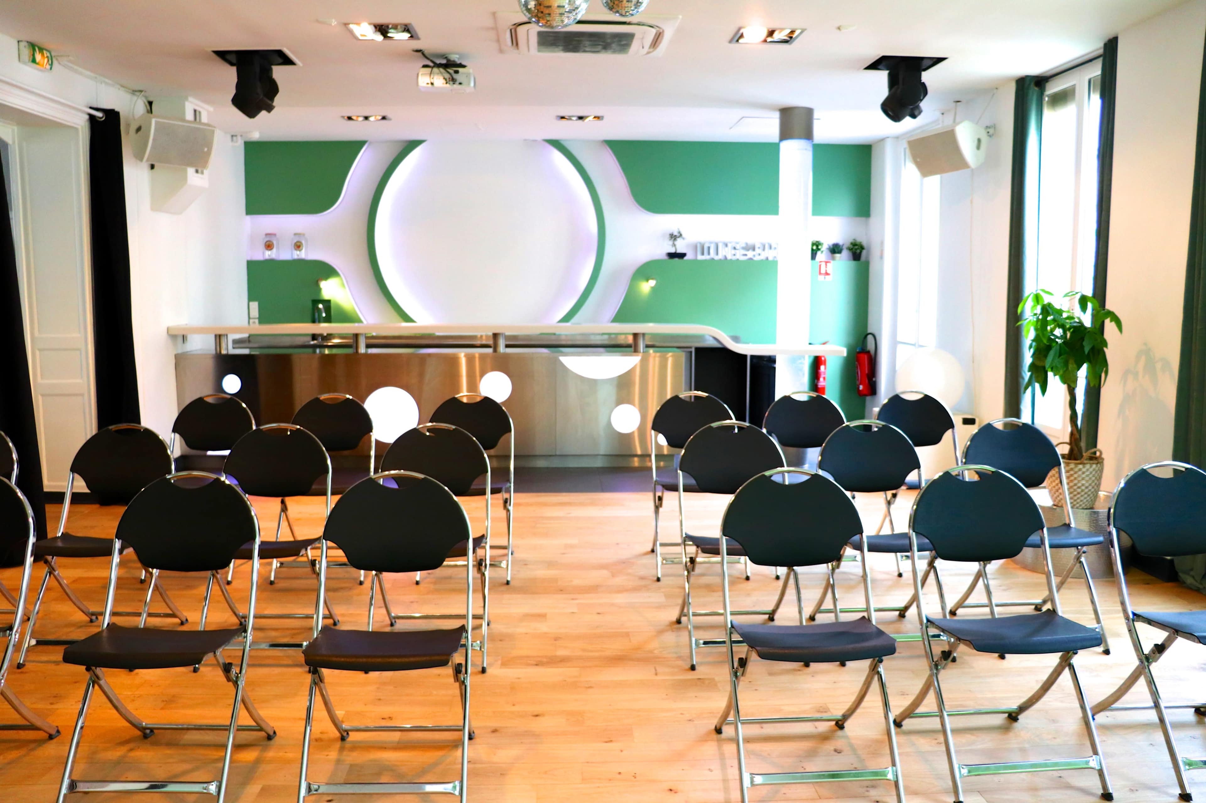 A modern event space features rows of empty black folding chairs facing a sleek reception area with green and white walls.