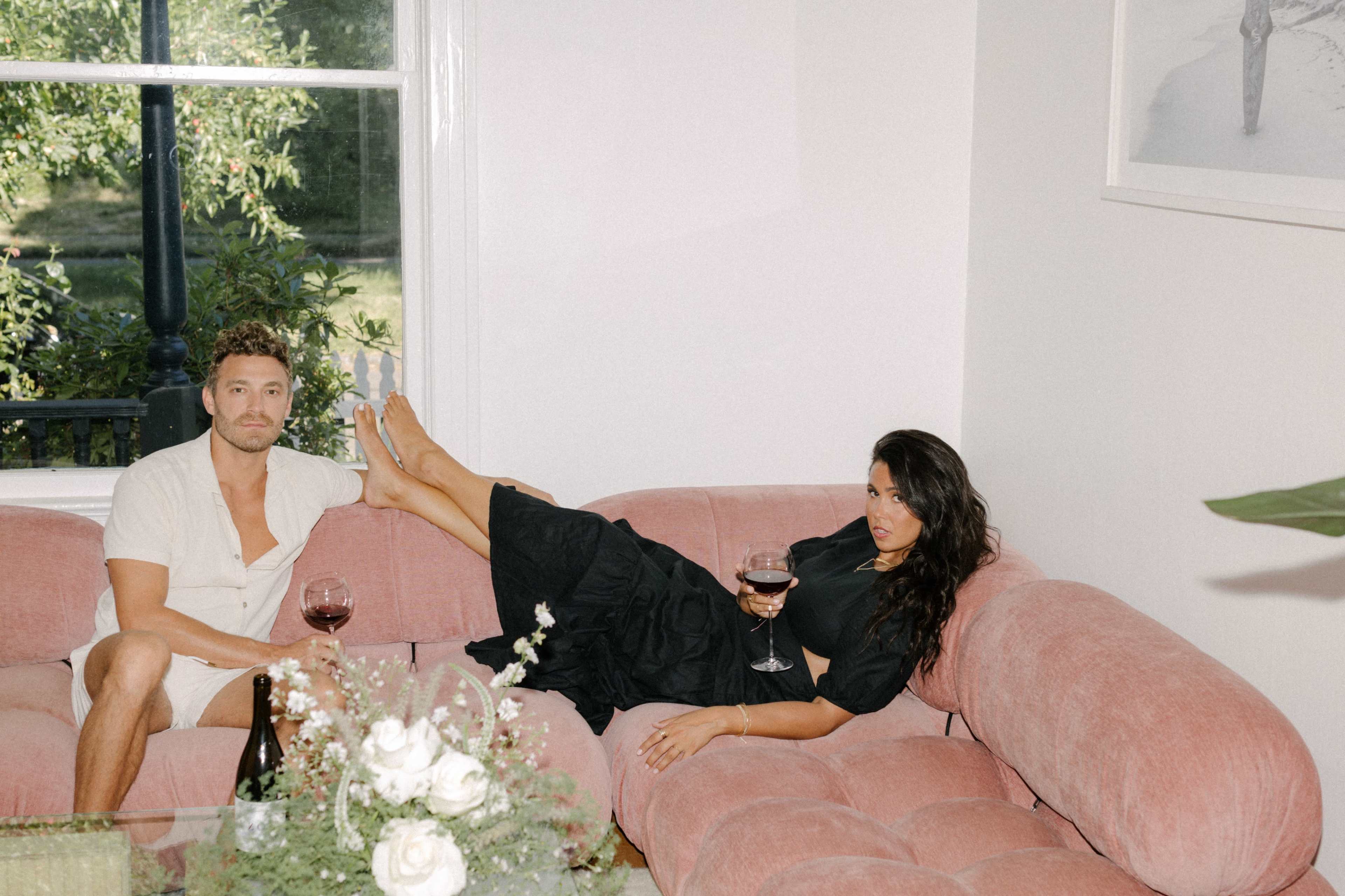 A man and a woman relax on a pink sofa in a well-lit lounge, each holding a glass of red wine.