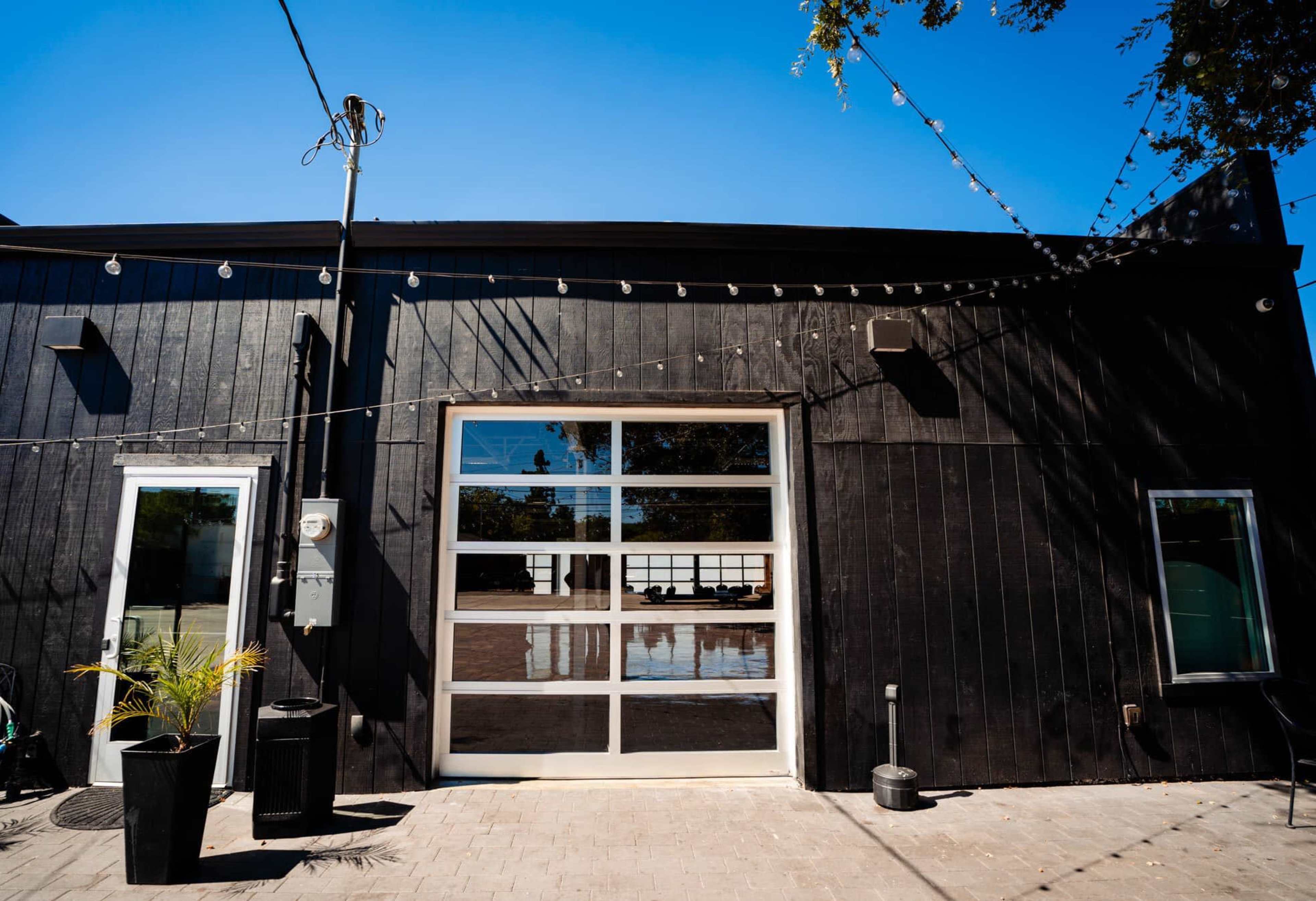 A modern black building features large glass garage doors and multiple windows, with string lights overhead and a clear blue sky in the background.