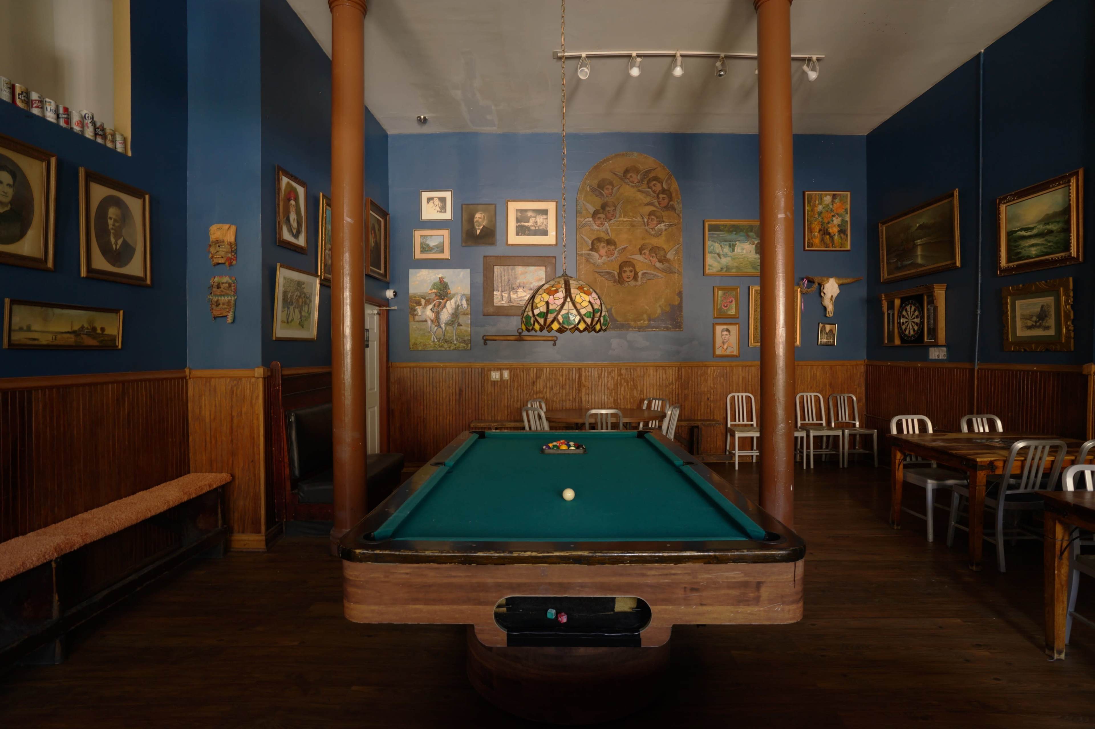 The image shows a billiard room featuring a green pool table surrounded by chairs and walls adorned with various framed artworks.