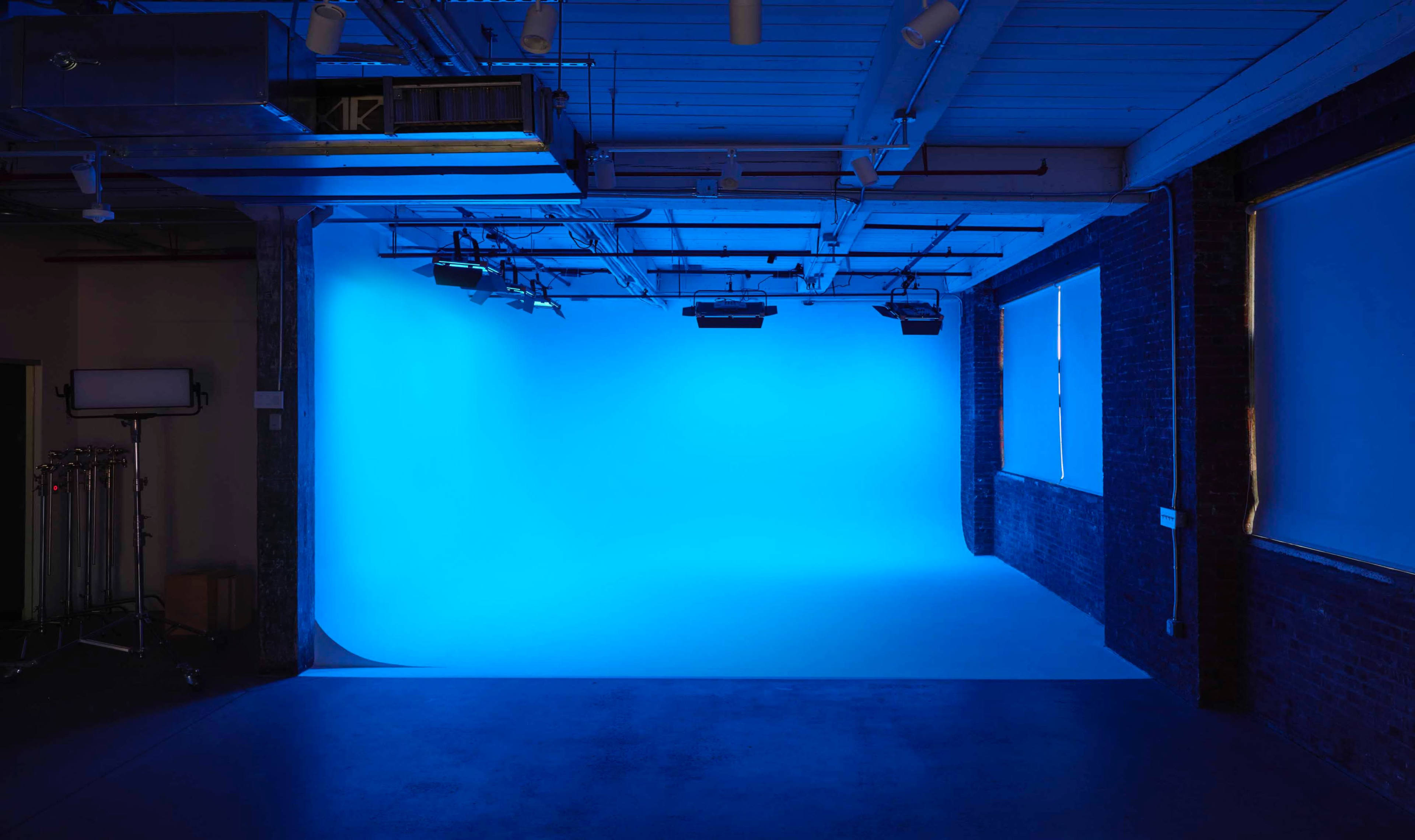 A photography studio featuring a blue backdrop, illuminated by overhead lights and natural light from a window.