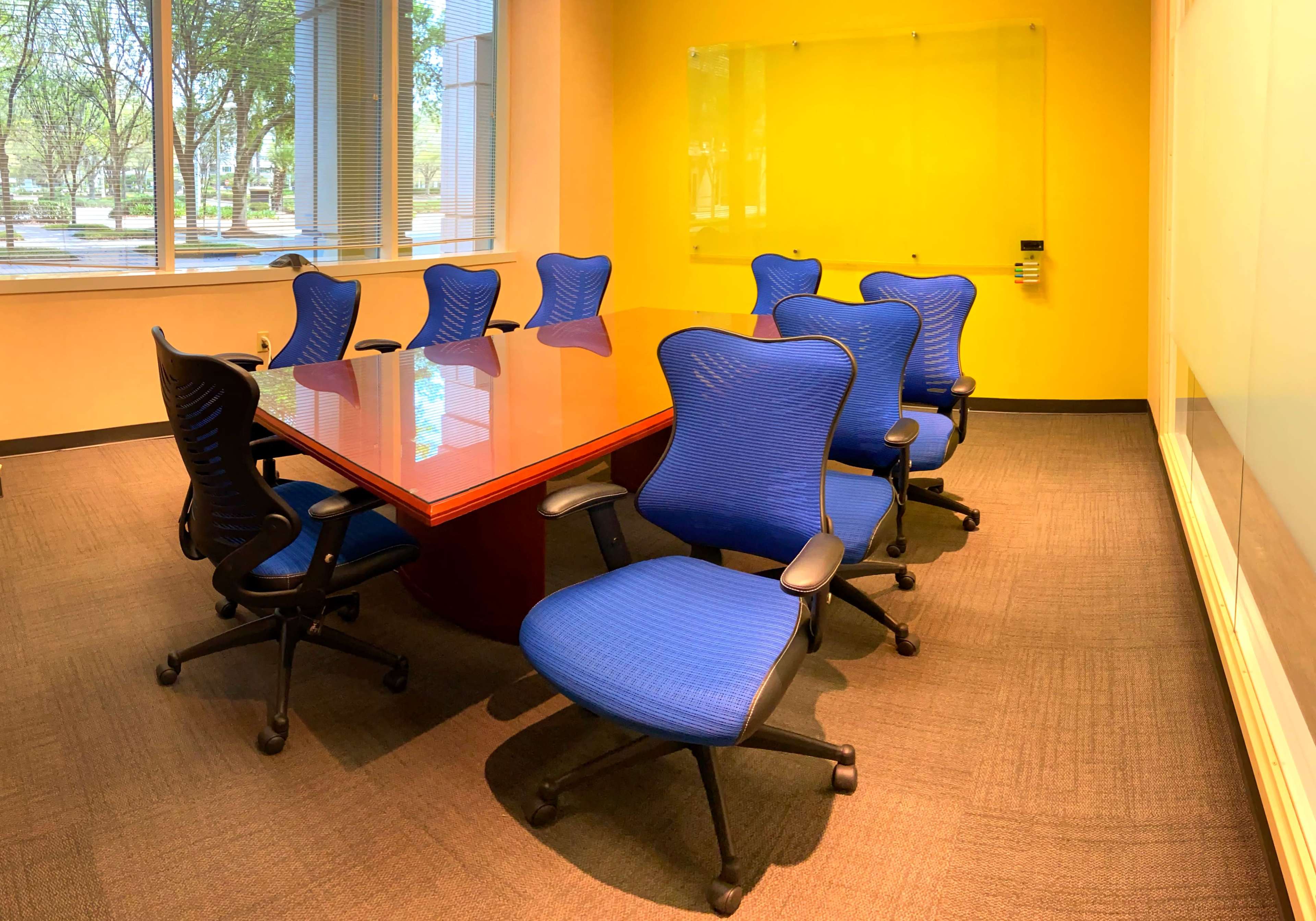 A conference room features a wooden table surrounded by eight blue ergonomic chairs against a bright yellow wall.