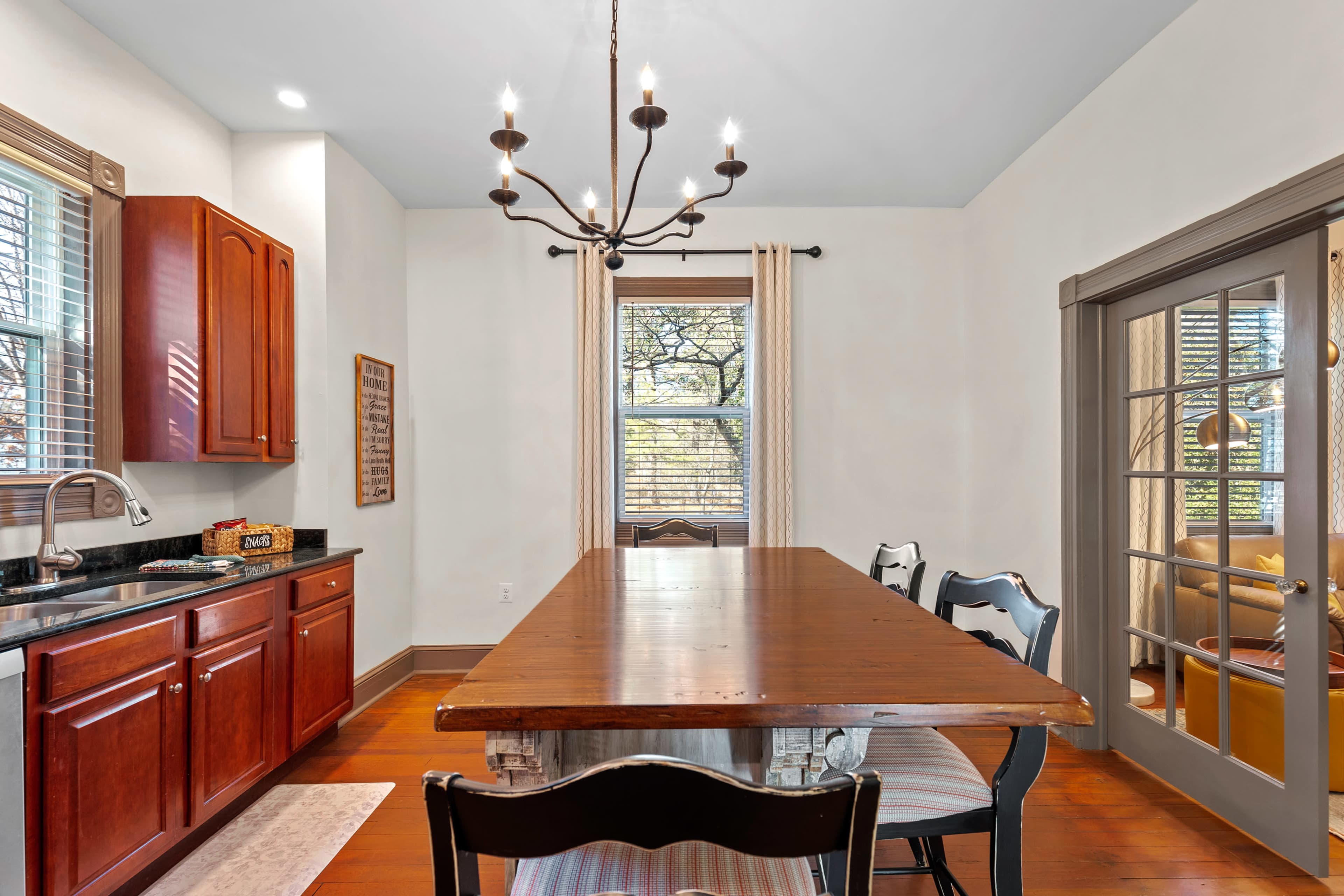A well-lit dining area features a long wooden table surrounded by chairs, with dark cabinets and a chandelier hanging above.