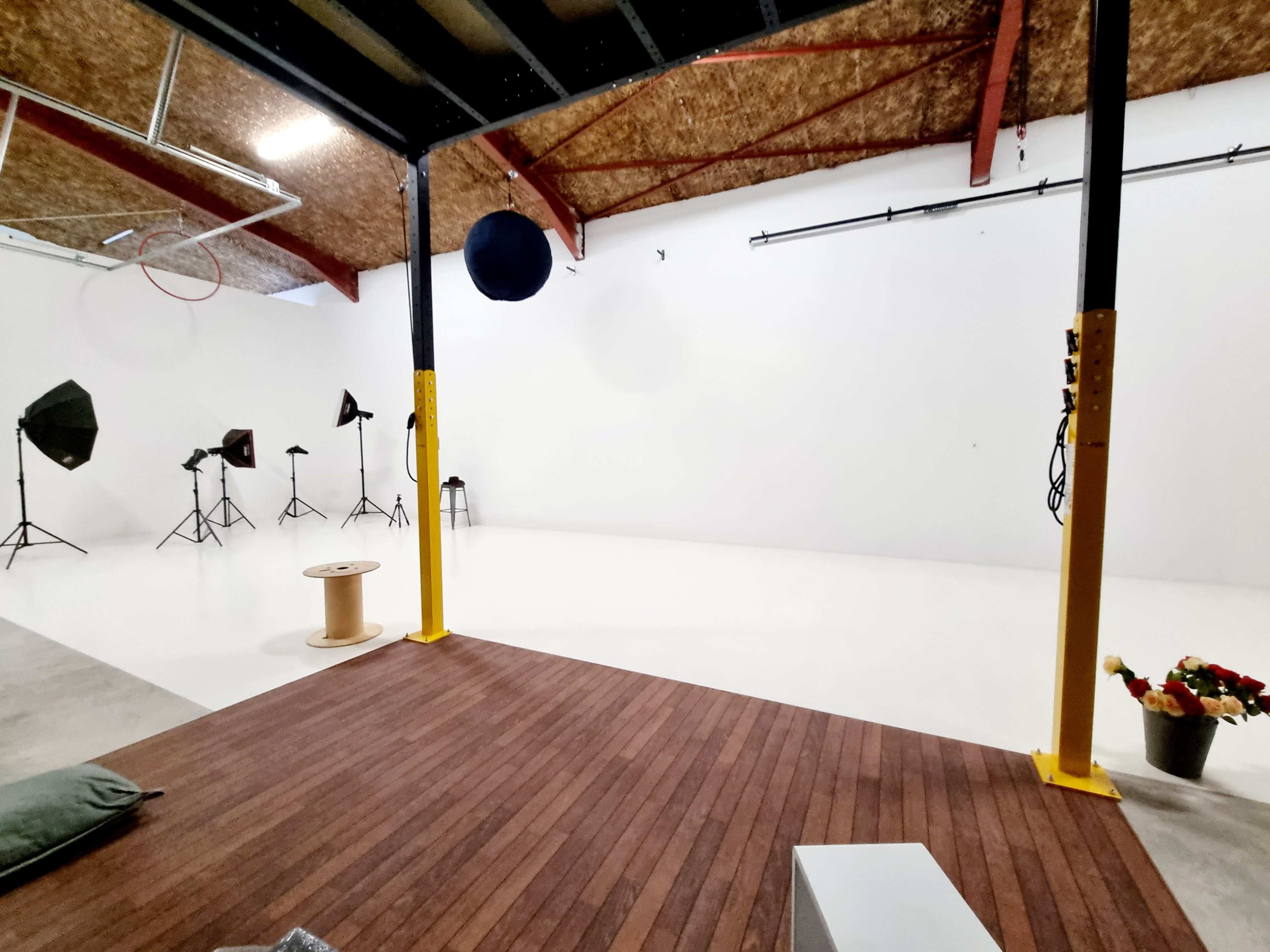 A spacious photography studio features a white backdrop, various lighting equipment on stands, and a wooden floor area with a small table and a flower arrangement.