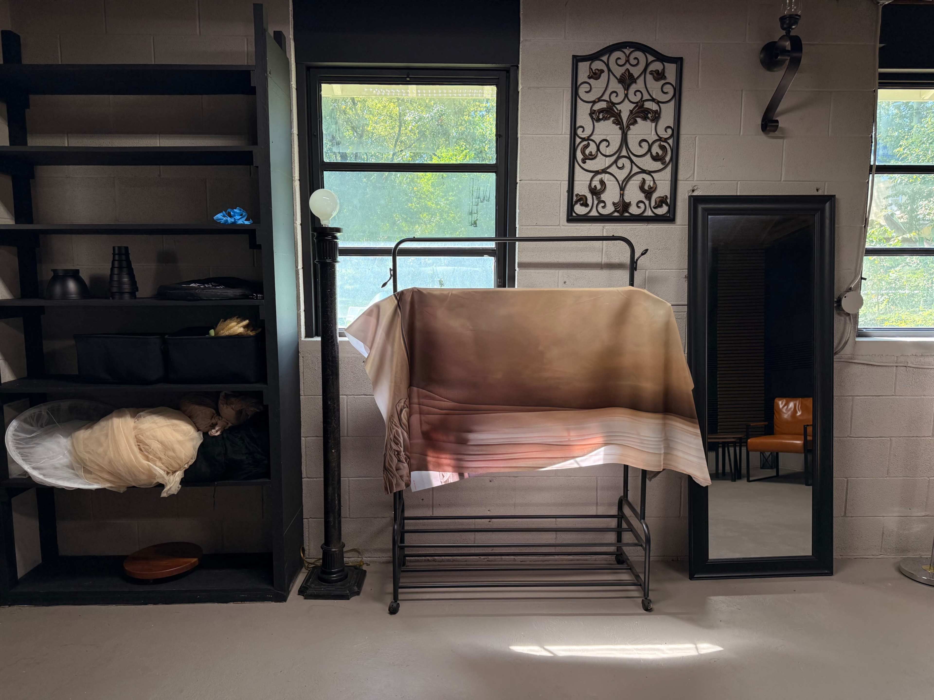 The image shows a room featuring a clothing rack with a blanket draped over it, a mirror, shelves with various items, and large windows allowing natural light to enter.