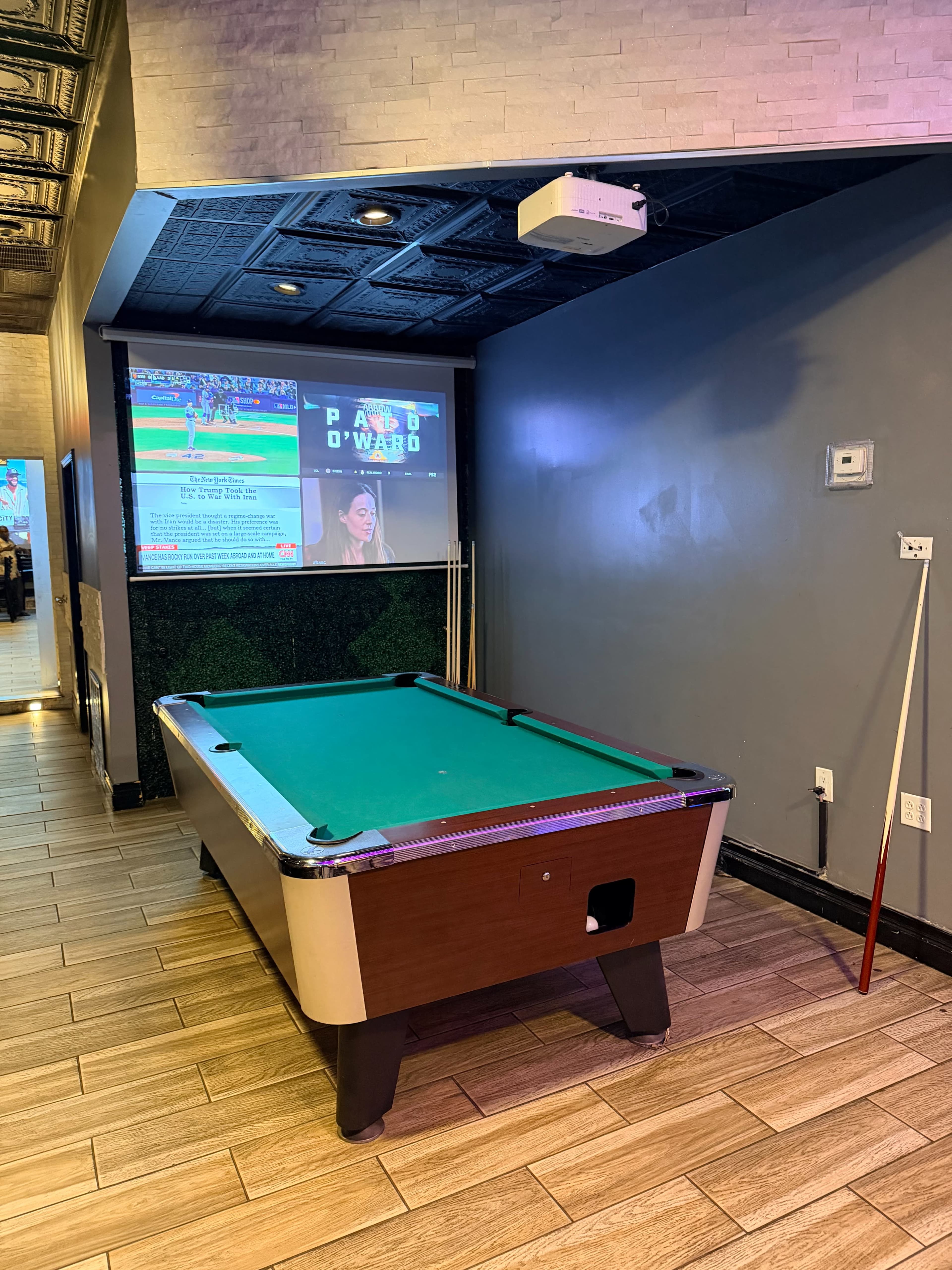 A pool table is positioned in a dimly lit room, with a projector screen displaying sports content on the wall behind it.