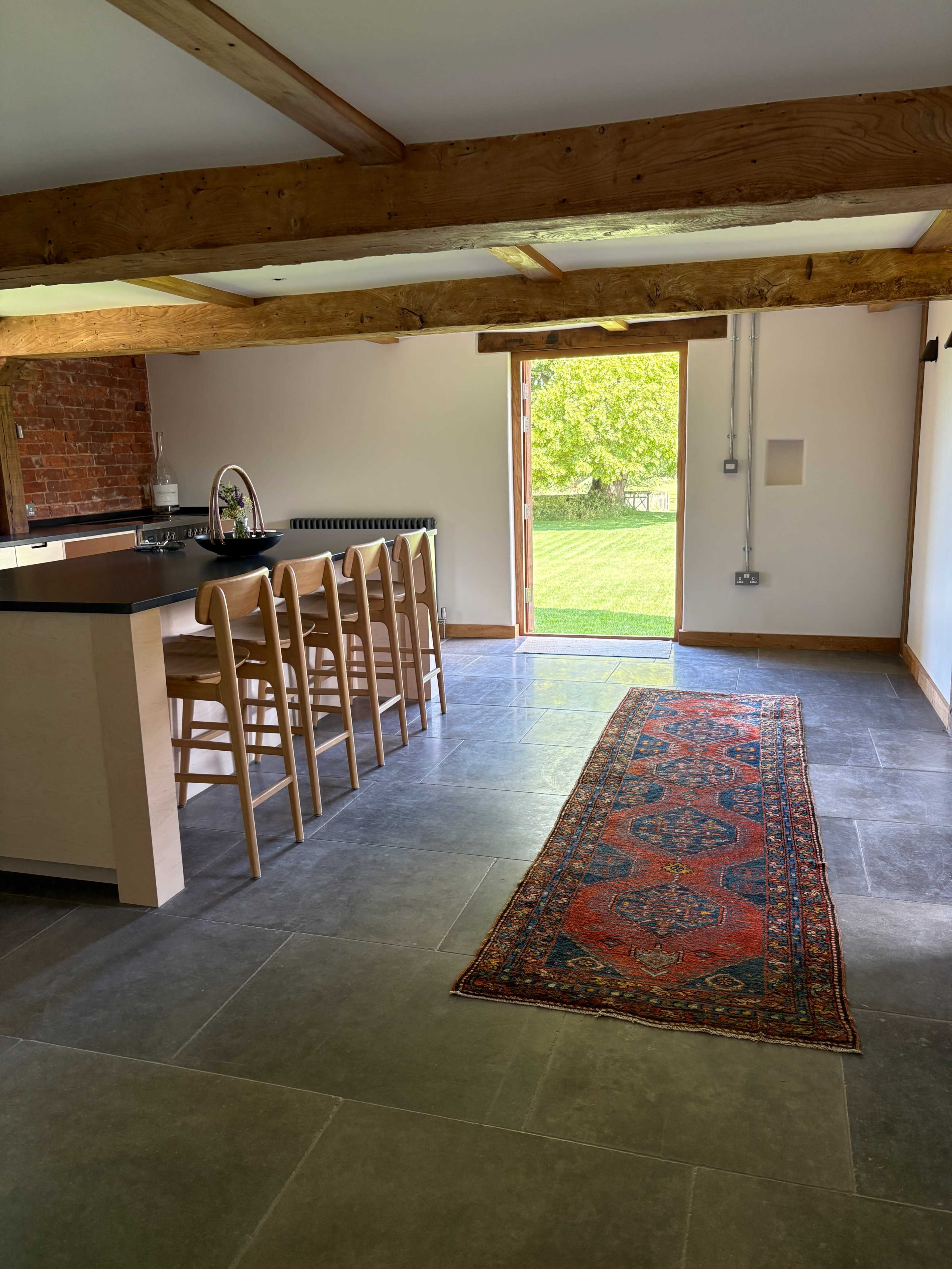 The image shows a modern kitchen area with wooden beams, a bar counter with stools, and a long rug leading to an open door that opens to a green outdoor space.