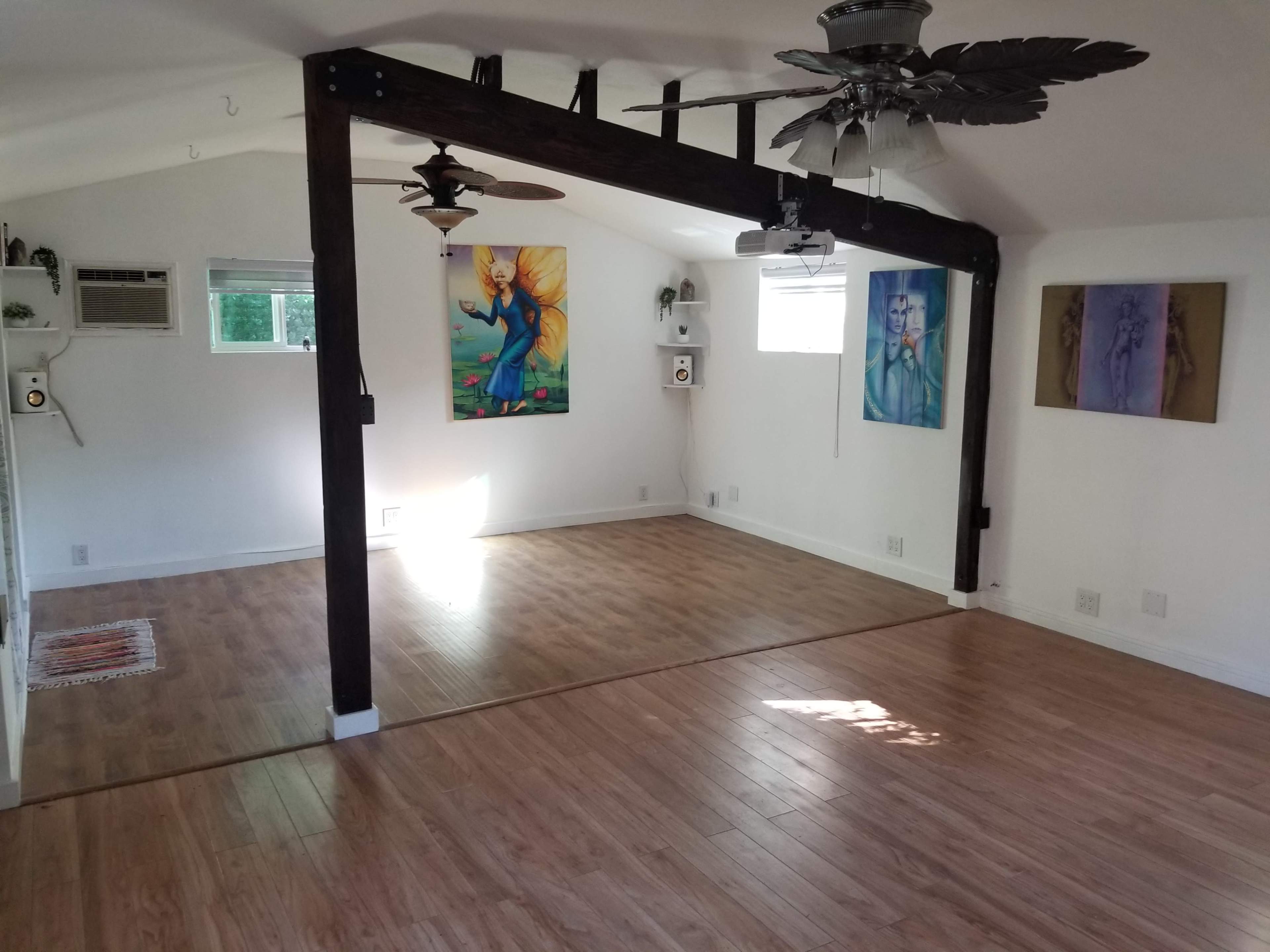 The image shows a spacious room with wooden flooring, featuring two decorative wall paintings, two ceiling fans, and a divided area created by a wooden beam.