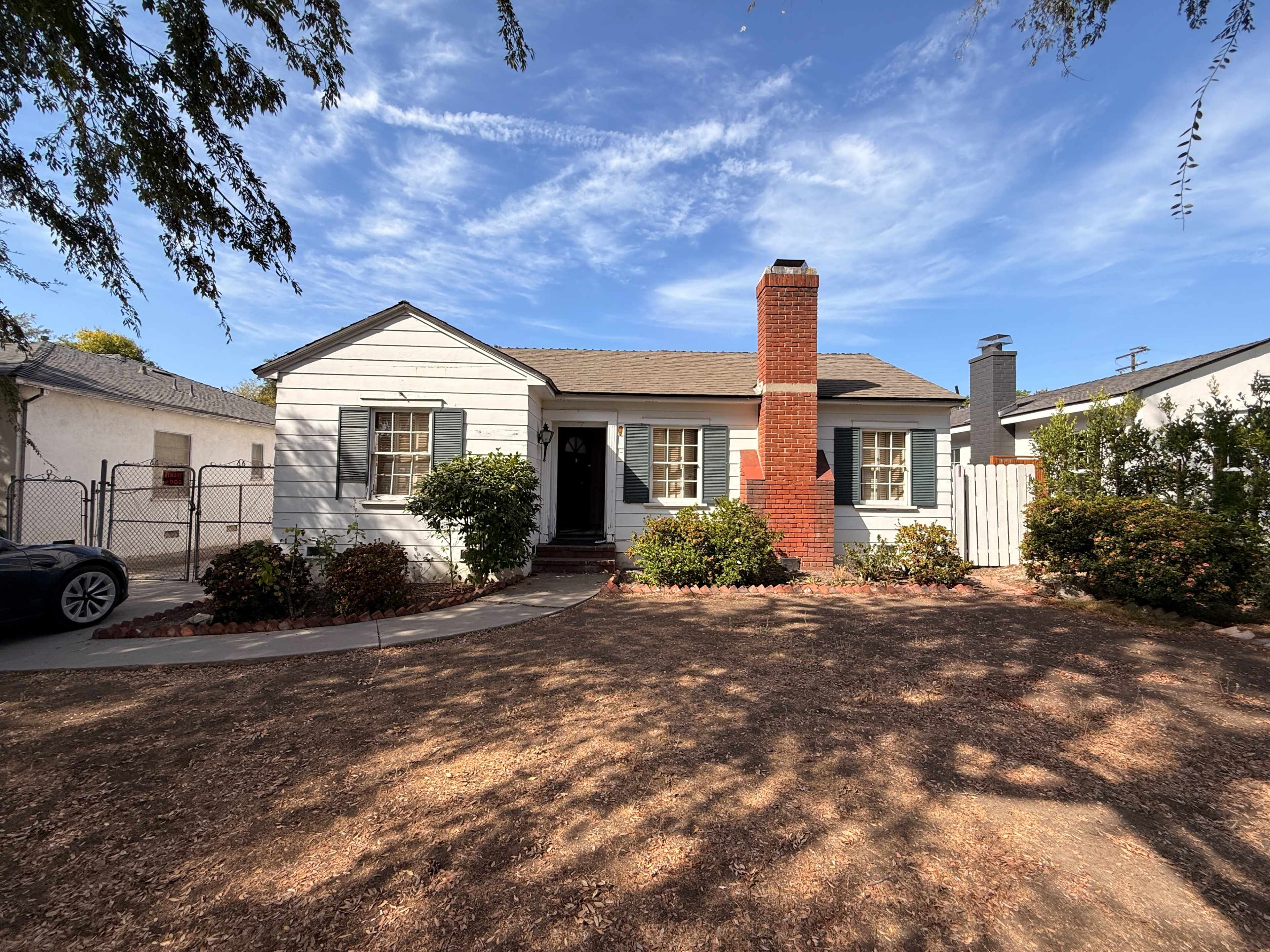 The image shows a single-story house with a red brick chimney, green shrubs, and a gravel front yard under a clear blue sky.