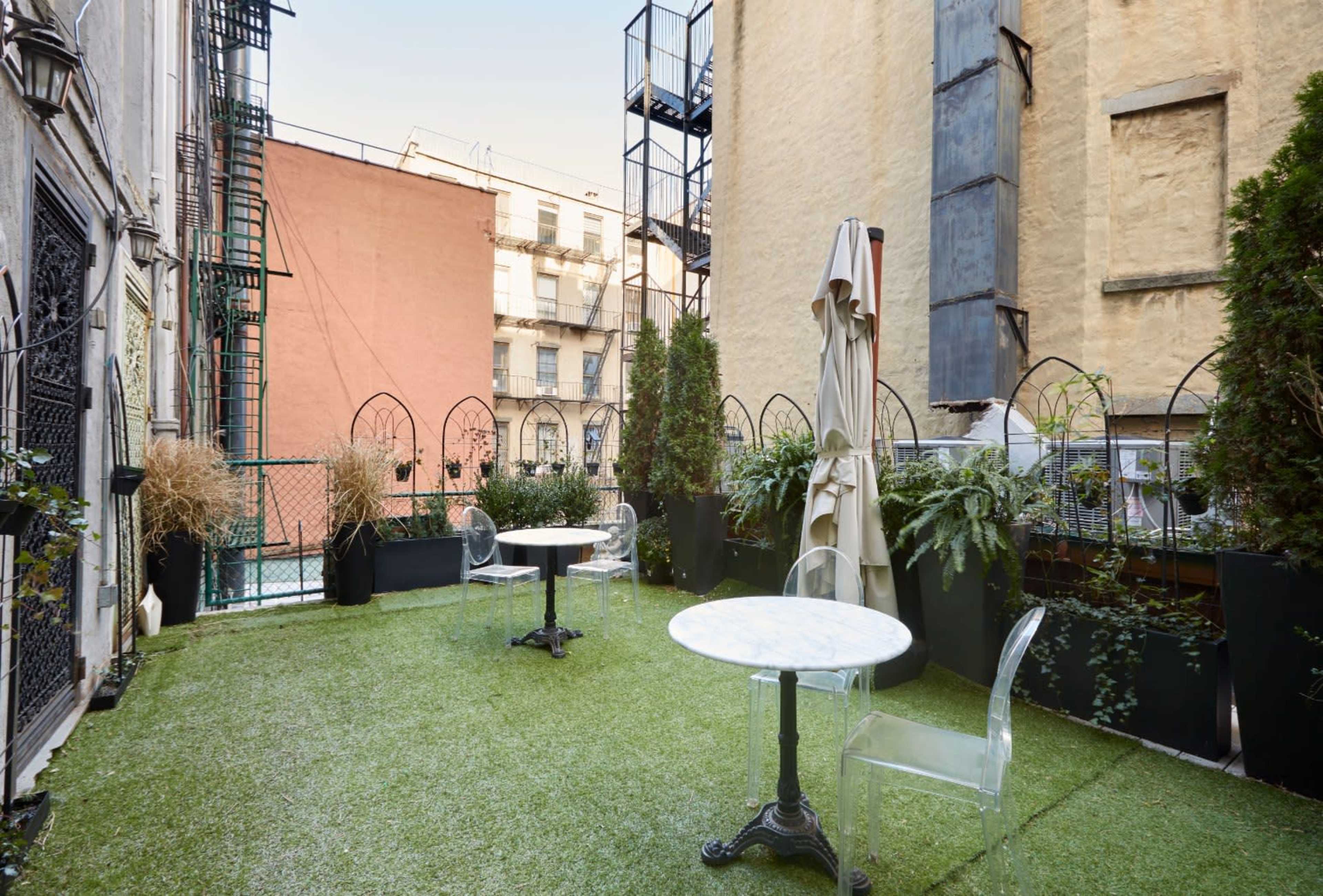 A small outdoor terrace with artificial grass, two marble-top tables, clear plastic chairs, and surrounding greenery in planters.