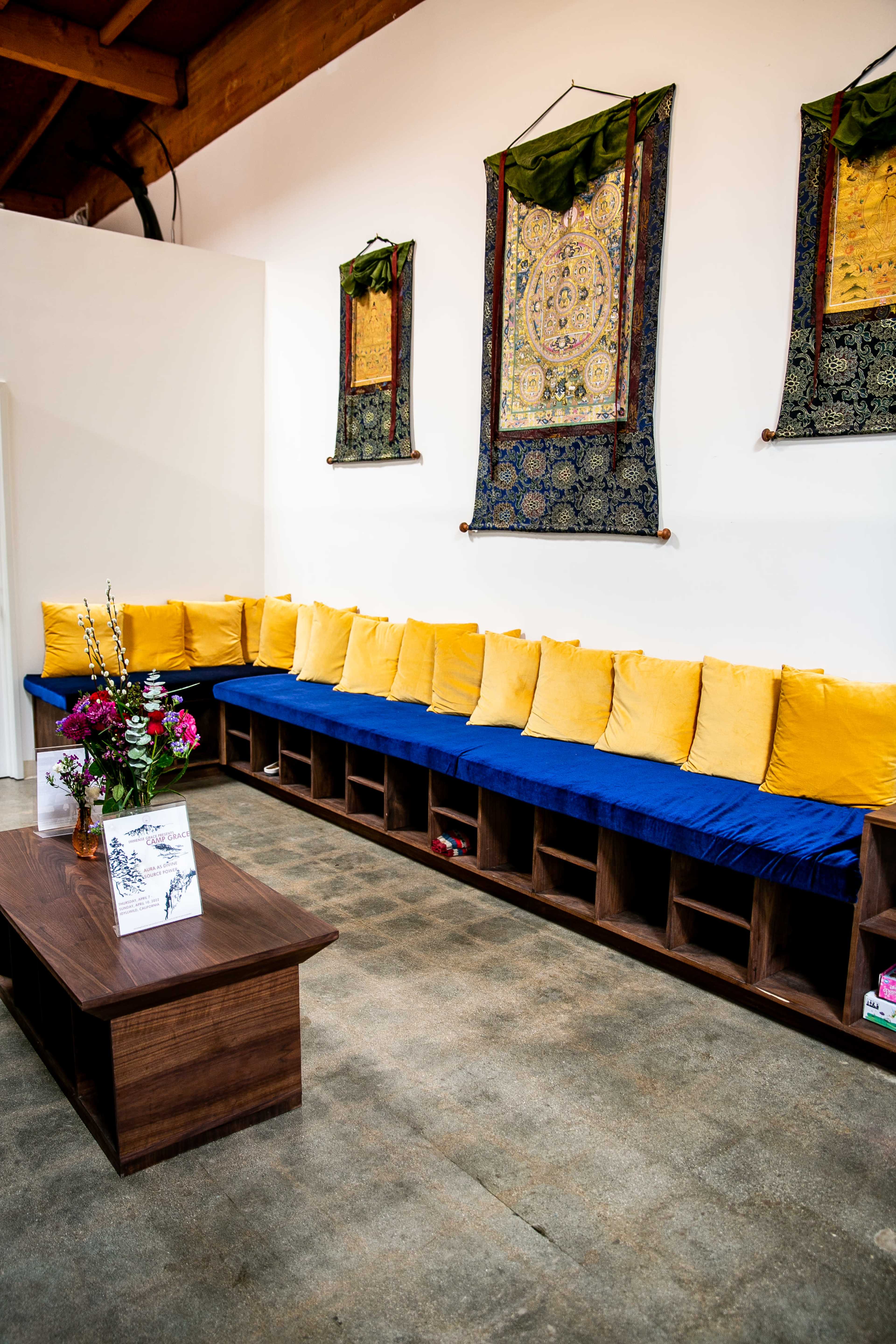 A cozy seating area features a long blue cushion-lined bench against a white wall, accompanied by a wooden coffee table and decorative wall hangings.