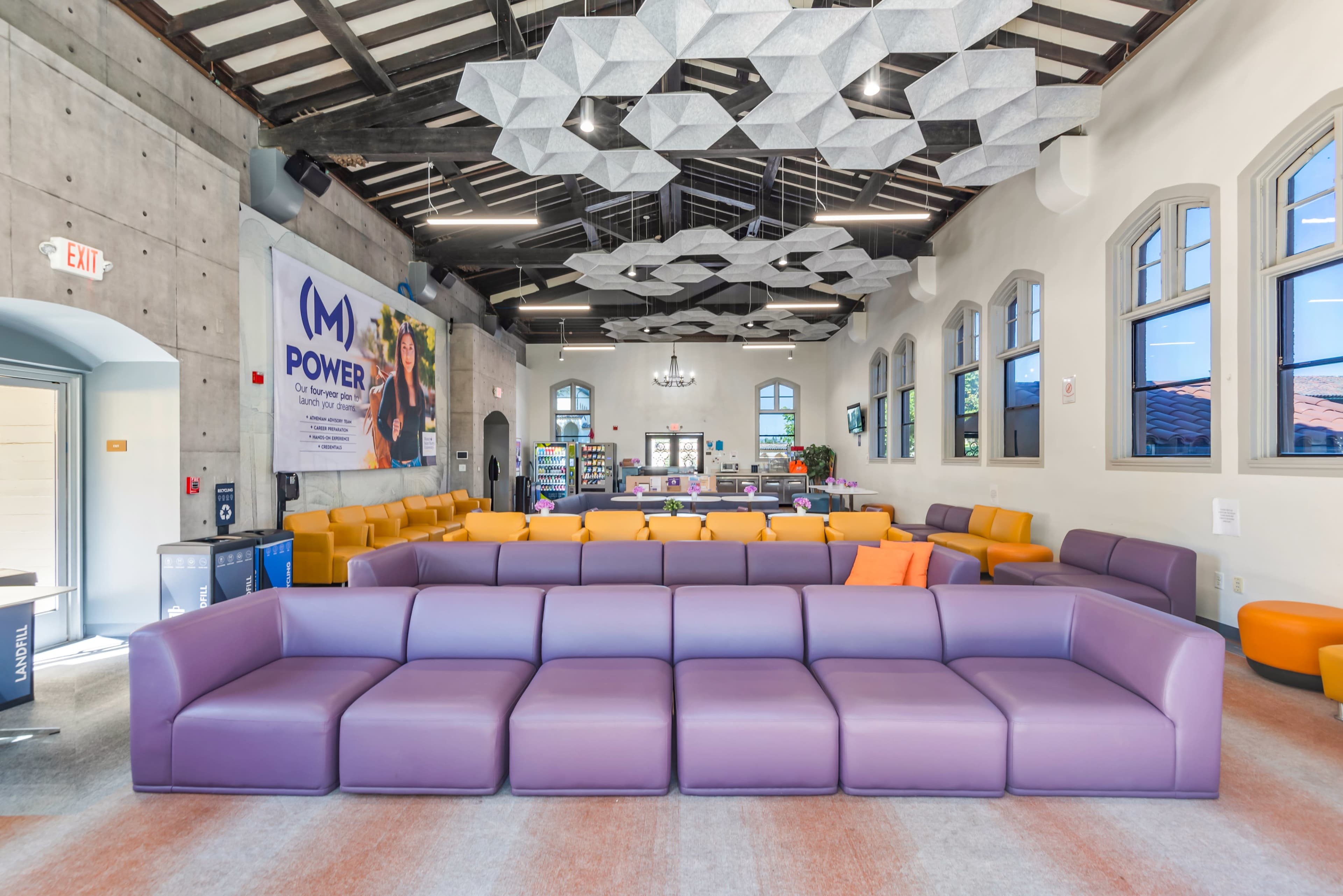 The image features a spacious lounge area with modern, colorful seating arrangements and unique ceiling installations.