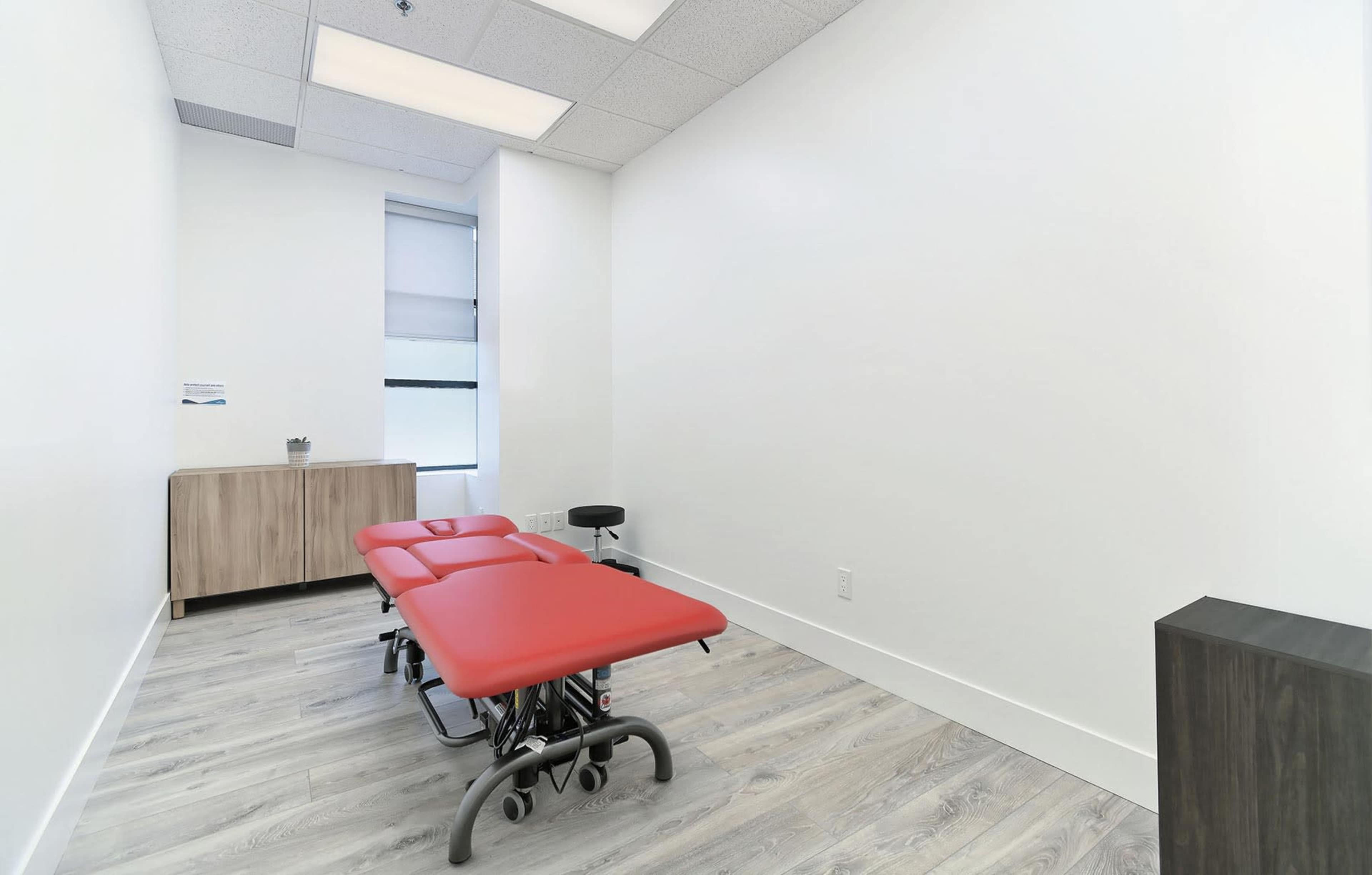 The image shows a minimalist treatment room featuring two red examination tables and a wooden cabinet against a white wall.