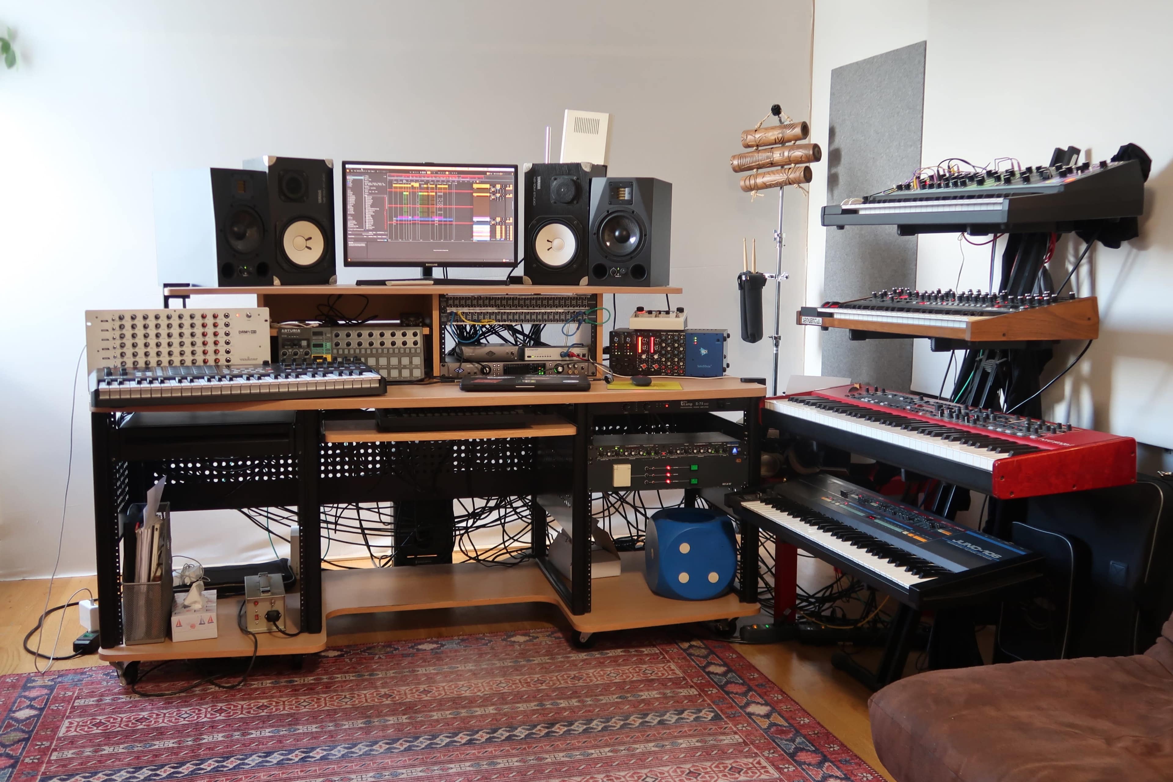 A music production workstation features a computer monitor, multiple speakers, synthesizers, a keyboard, and various equipment organized on a wooden desk.