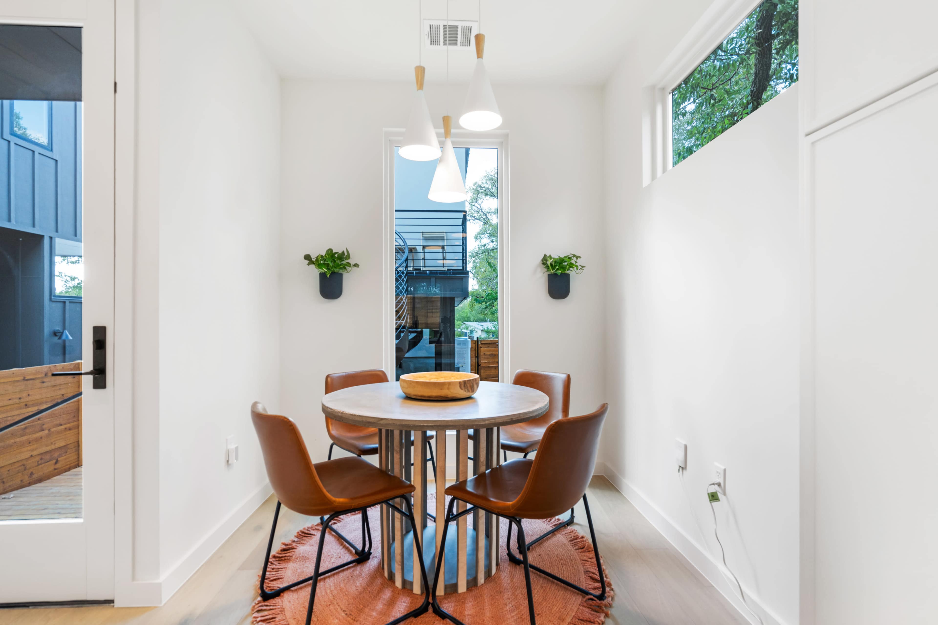 A round dining table with four chairs is positioned in a bright room featuring large windows and plant accents.
