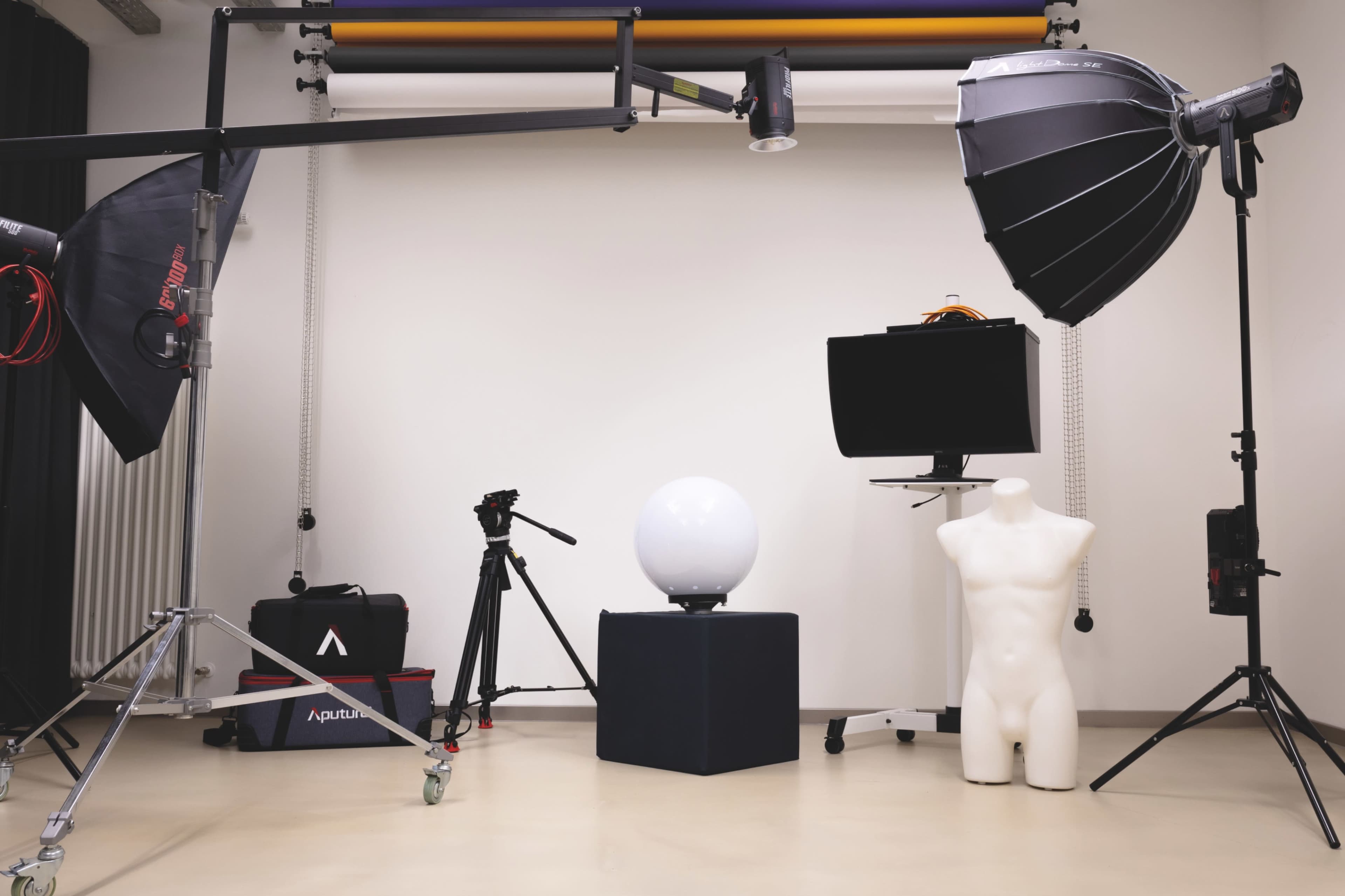 The image shows a photography studio setup with lighting equipment, a tripod, a mannequin, a spherical light on a pedestal, and a monitor against a plain wall.