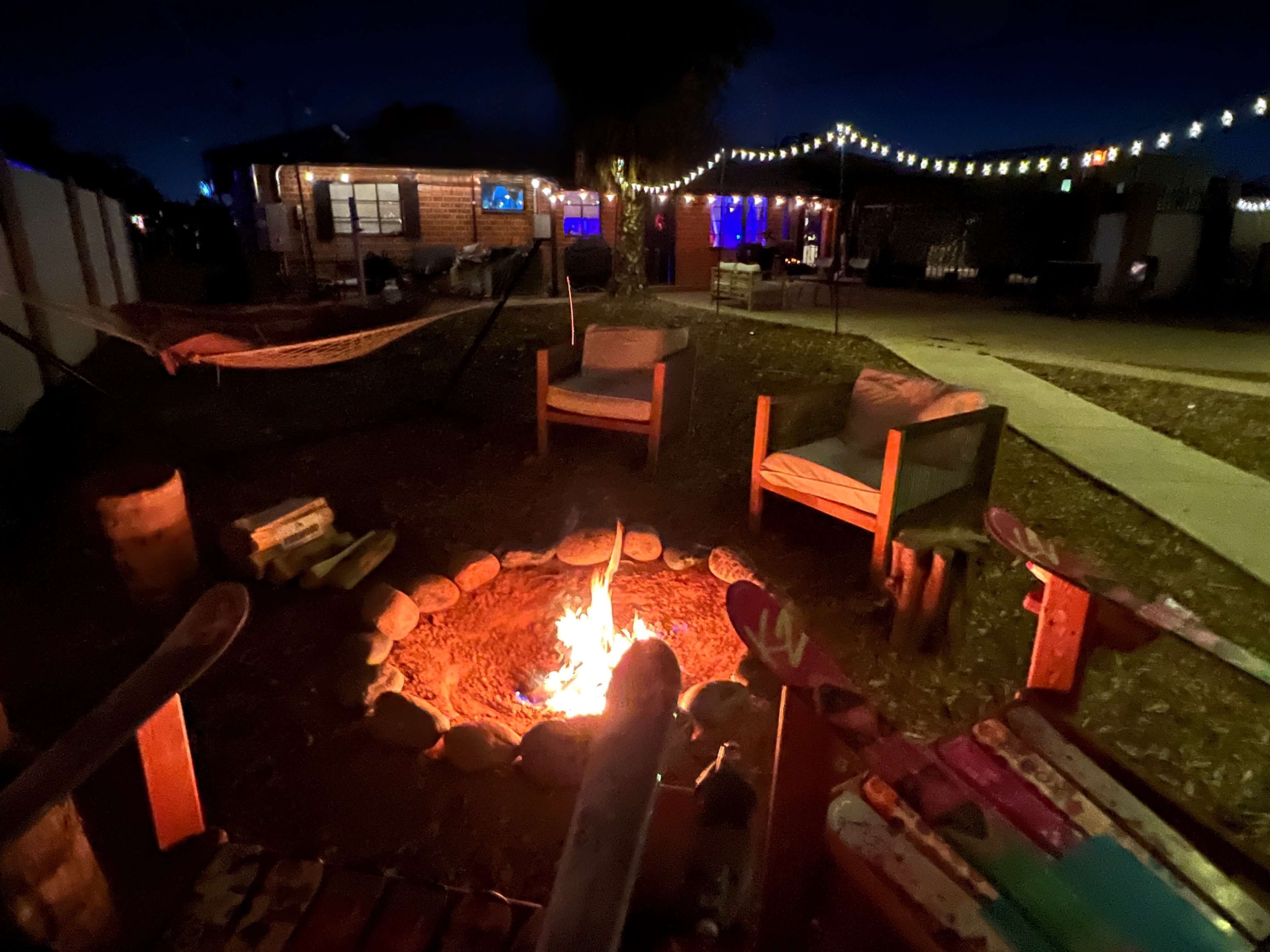 A campfire surrounded by wooden chairs and a rustic outdoor setting is illuminated by string lights at night.