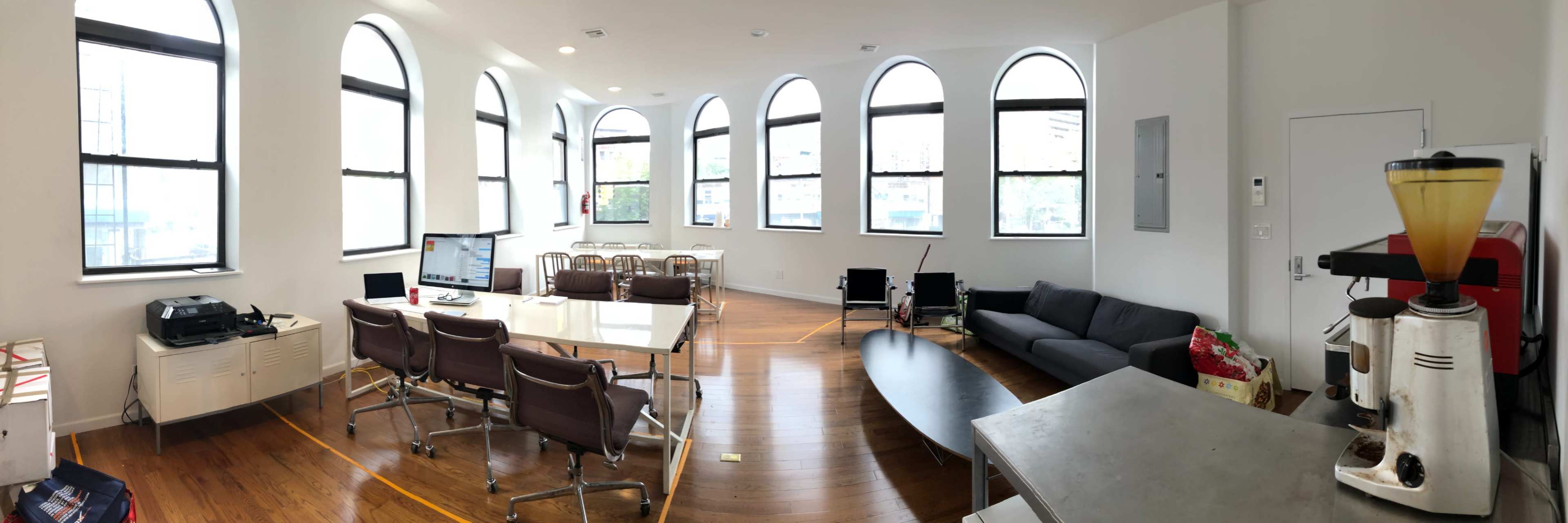 The image shows a modern office space with large arched windows, a conference table surrounded by chairs, a sofa, and a coffee machine in the corner.