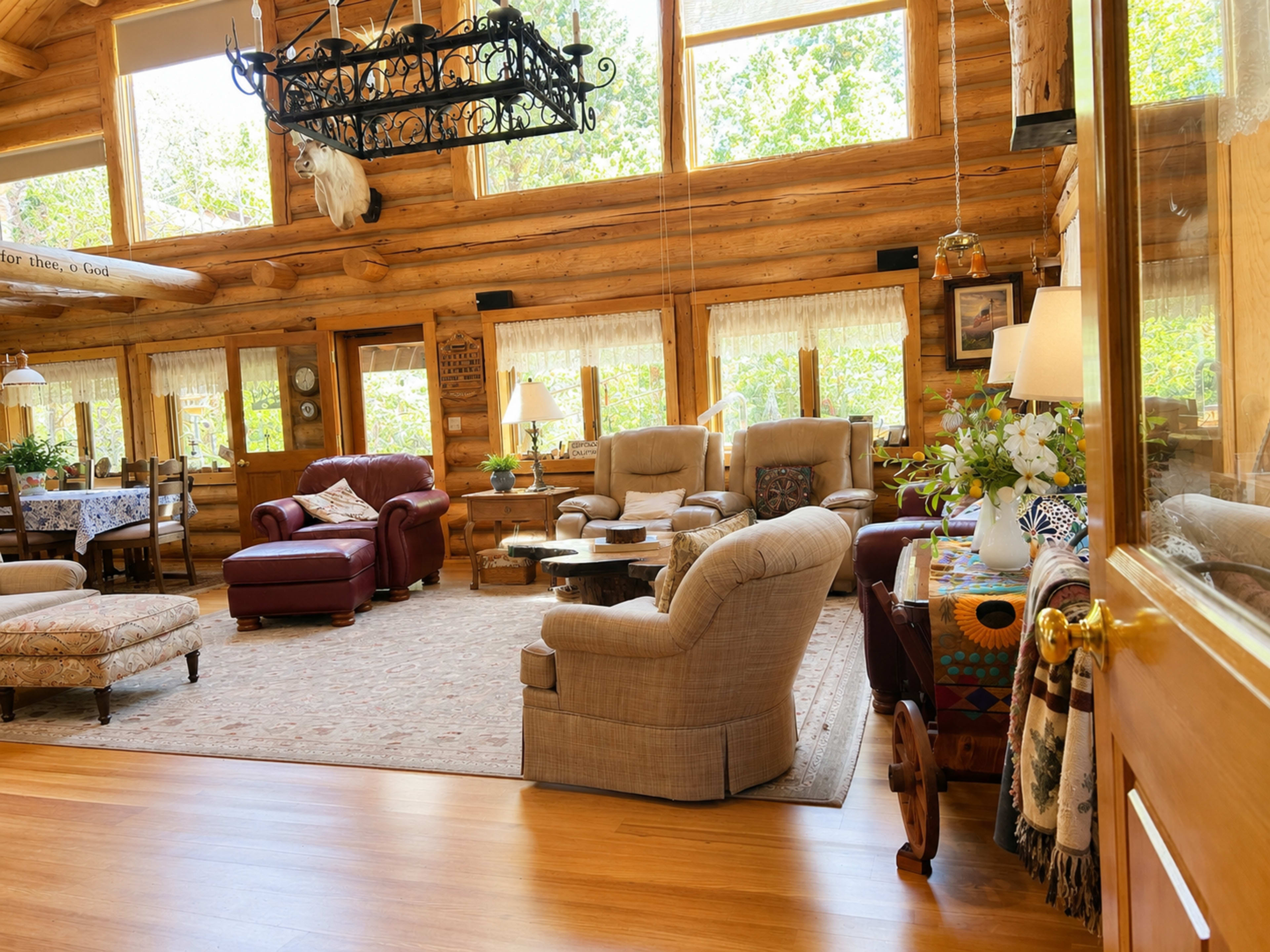 The image shows a cozy, rustic living room with wooden walls, large windows, and comfortable seating arranged around a central area rug.