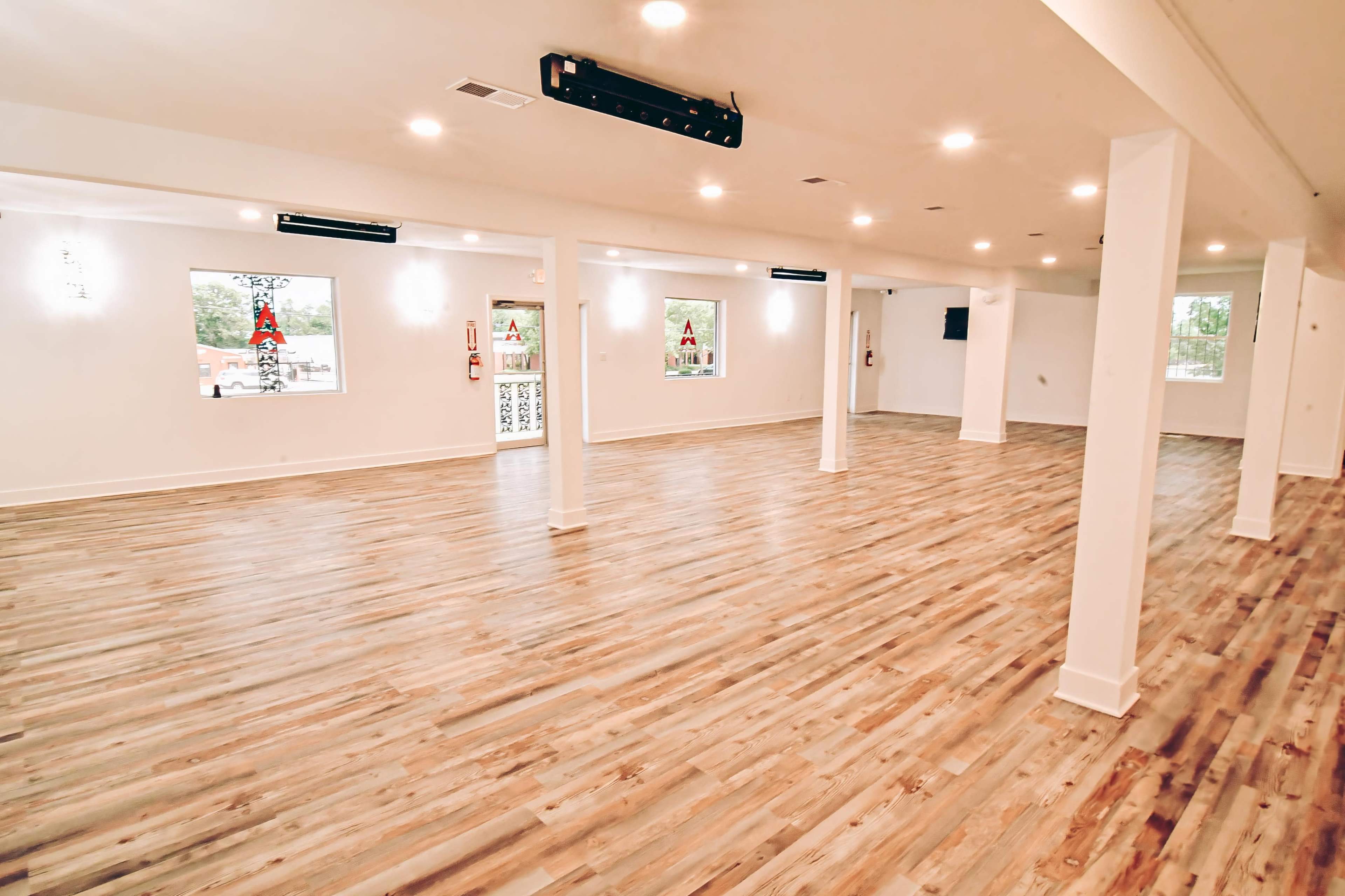 The image shows a spacious interior with wooden flooring, bright overhead lights, and several columns, creating an open and minimalist design.