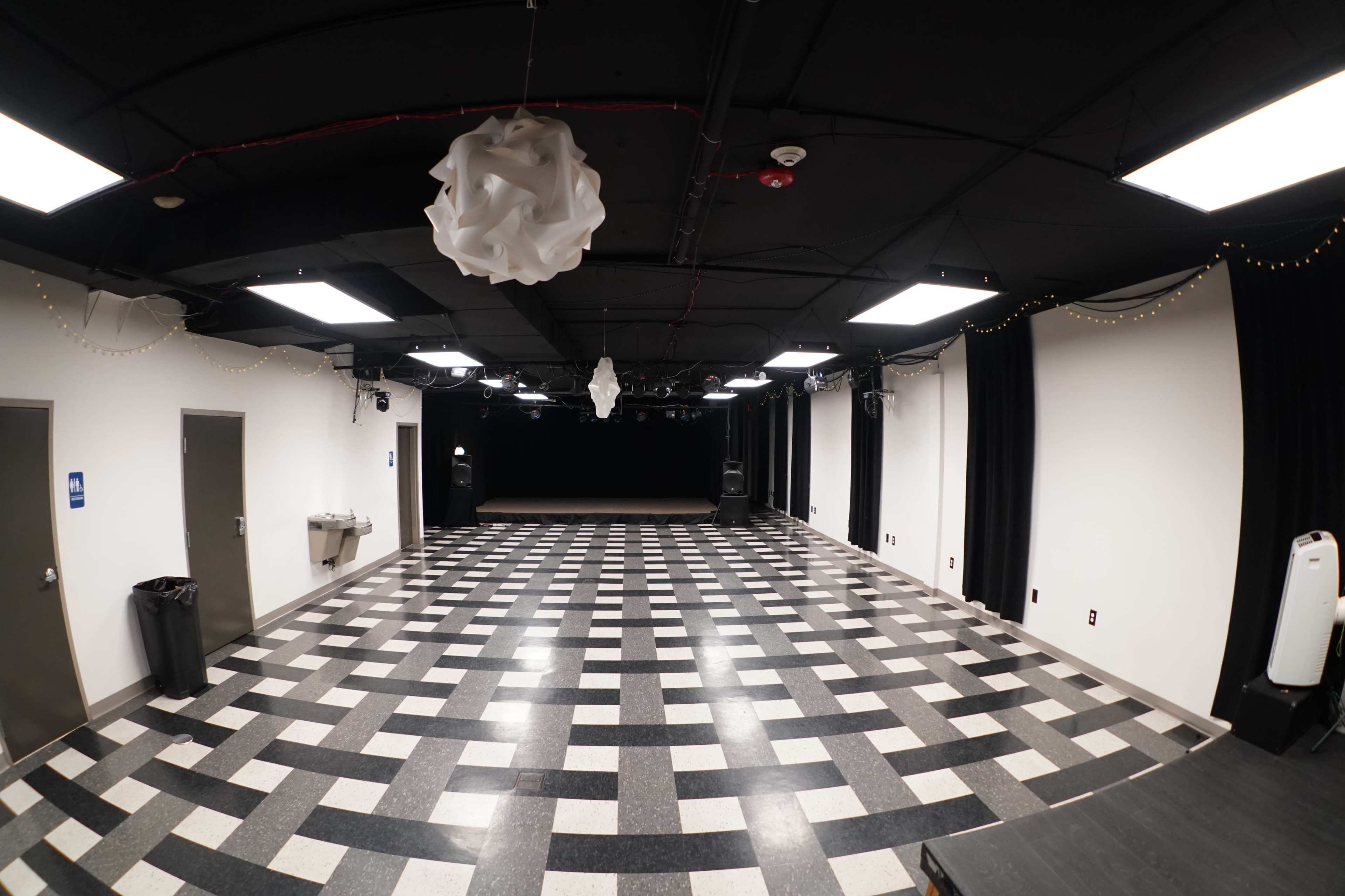 A spacious, empty room with a black and white checkered floor, black curtains, and bright overhead lights.