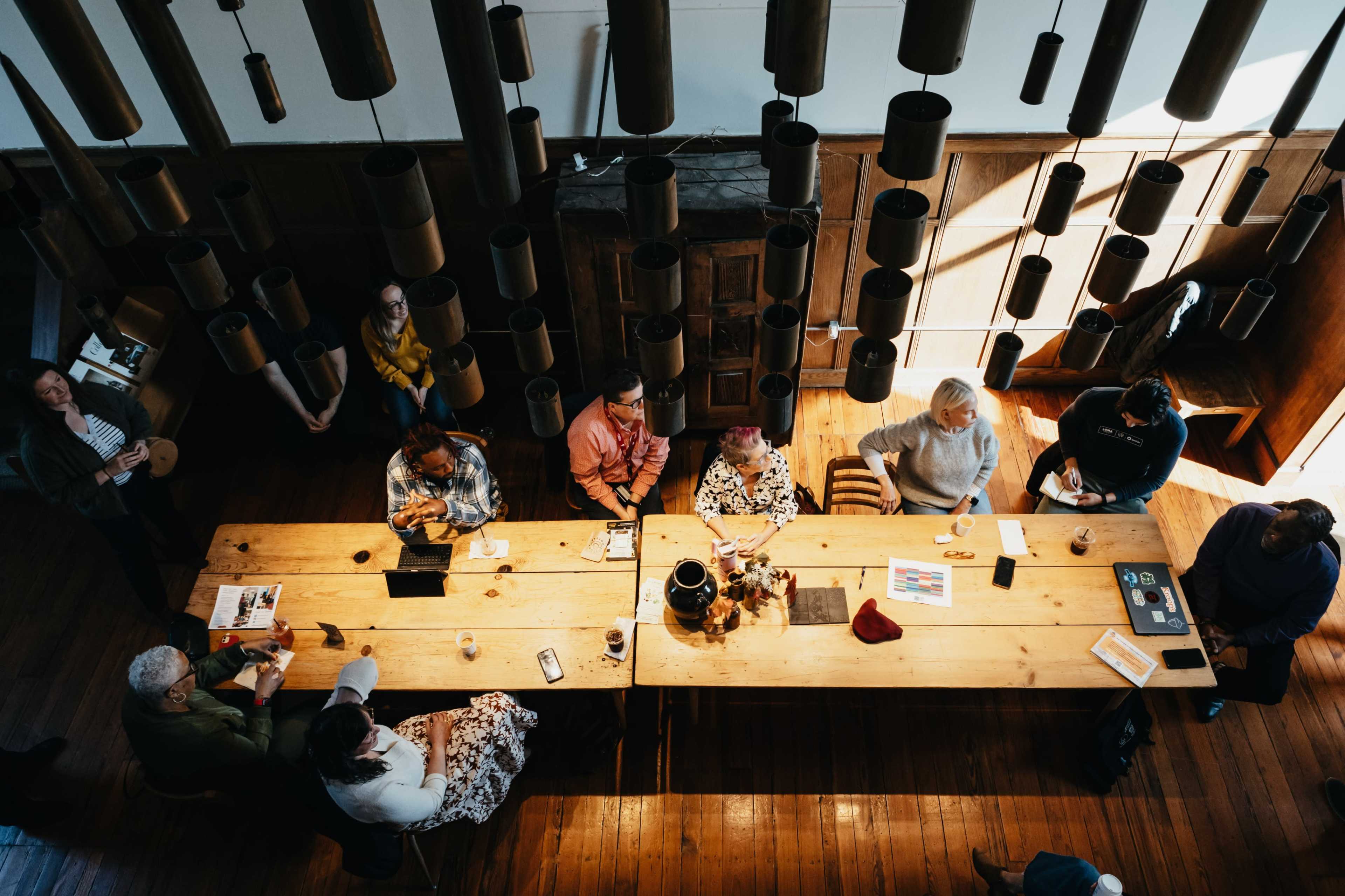 A group of people sits at a long wooden table in a well-lit room with hanging cylindrical lamps.
