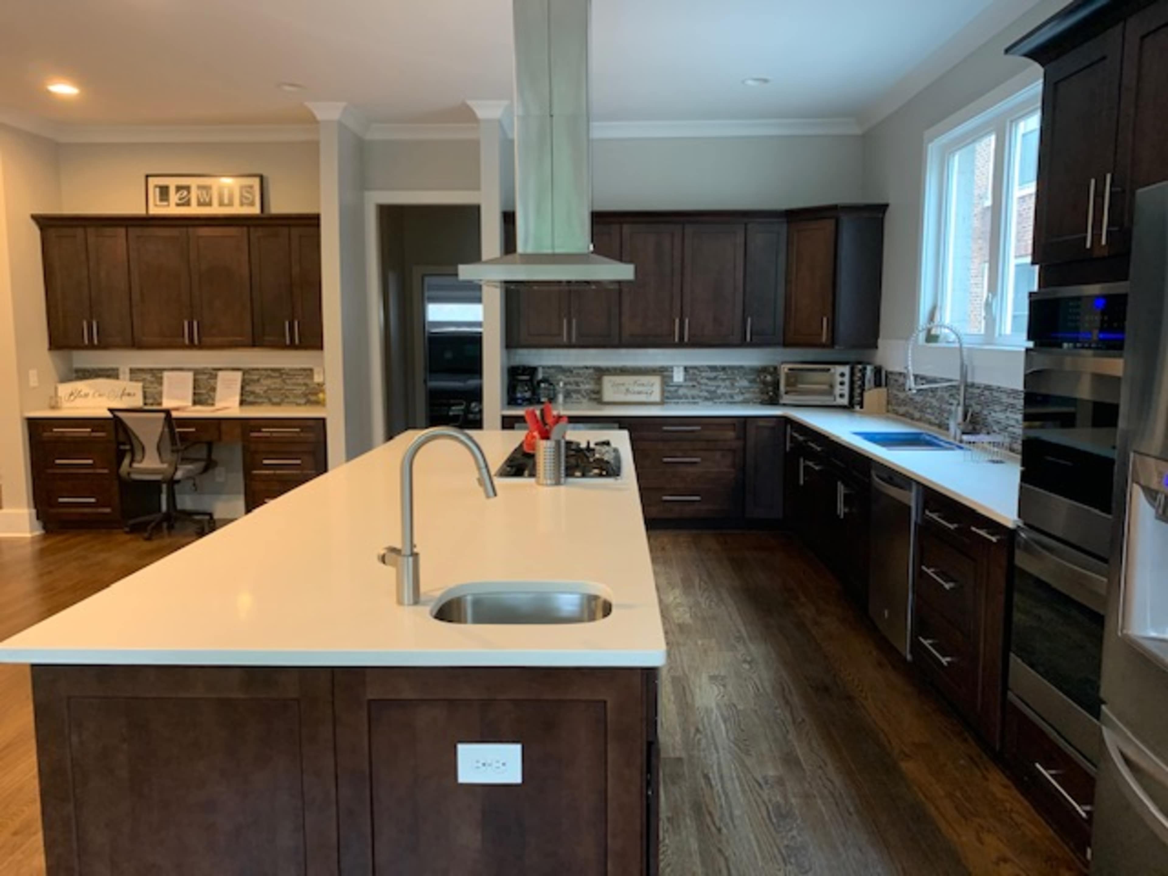 The image shows a modern kitchen with dark wood cabinets, a central island with a sink, and stainless steel appliances, illuminated by natural light from the windows.