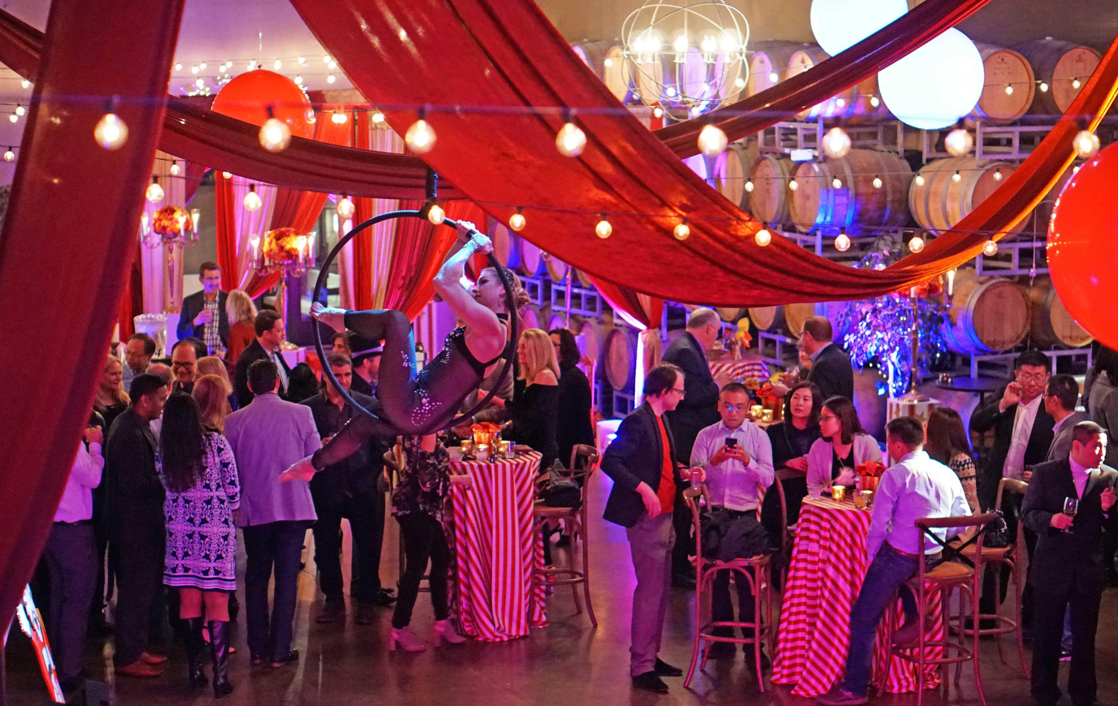 Aerial performer entertains guests at a lively event held in a venue adorned with red drapes and illuminated by string lights.