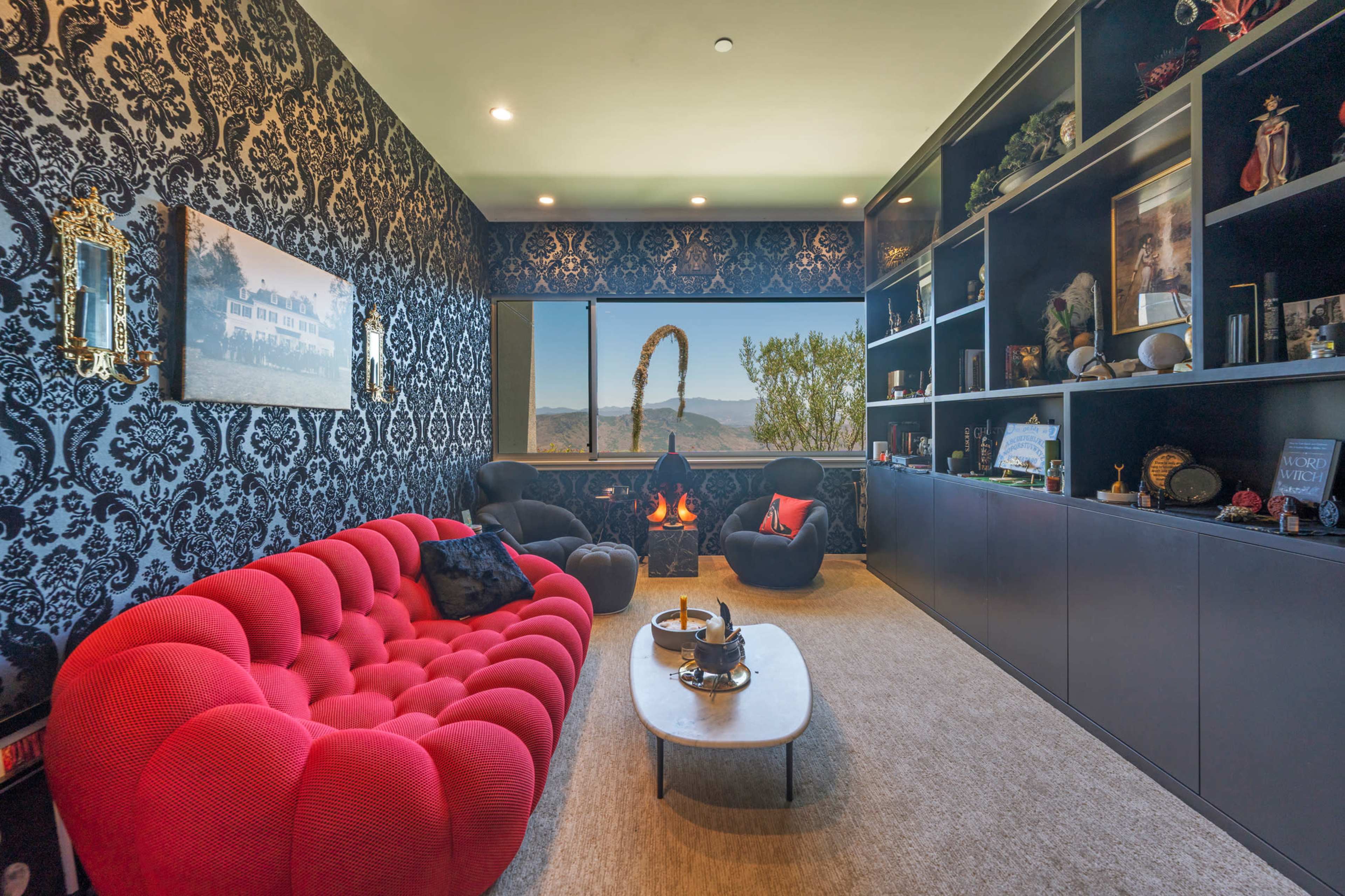 The image shows a modern living room with a red tufted sofa, a black wall adorned with intricate wallpaper, and a large window displaying a scenic view of mountains.