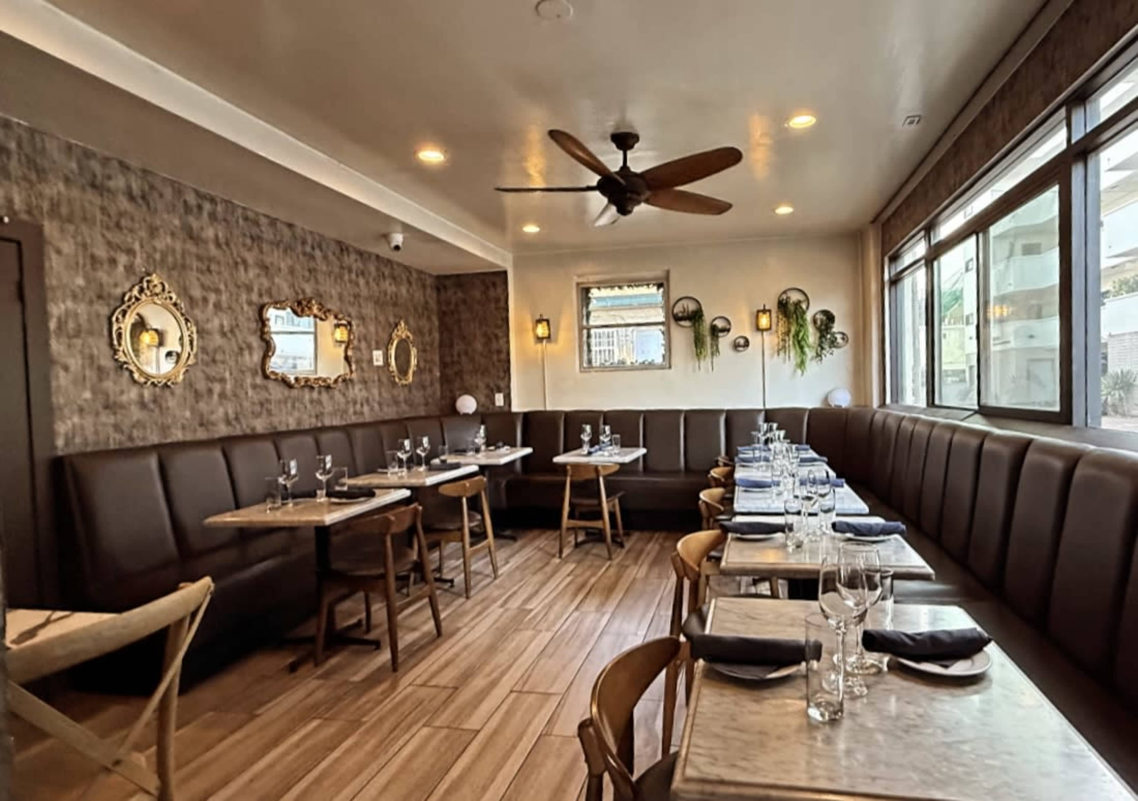 The image shows a modern restaurant interior featuring brown leather seating, round tables with white tablecloths, and large windows allowing natural light.