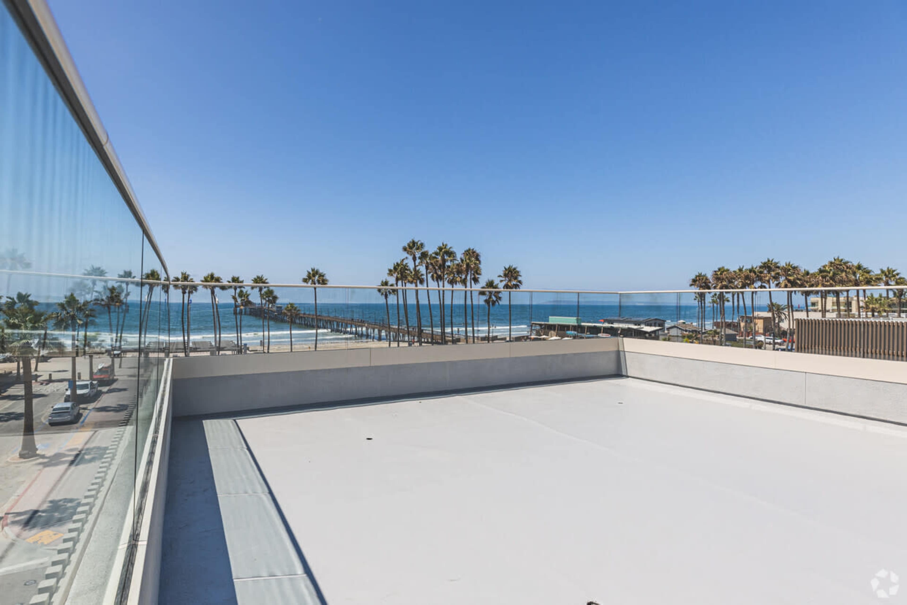 The image shows a view from a rooftop terrace overlooking a beach, with palm trees lining the shore and a pier extending into the ocean.