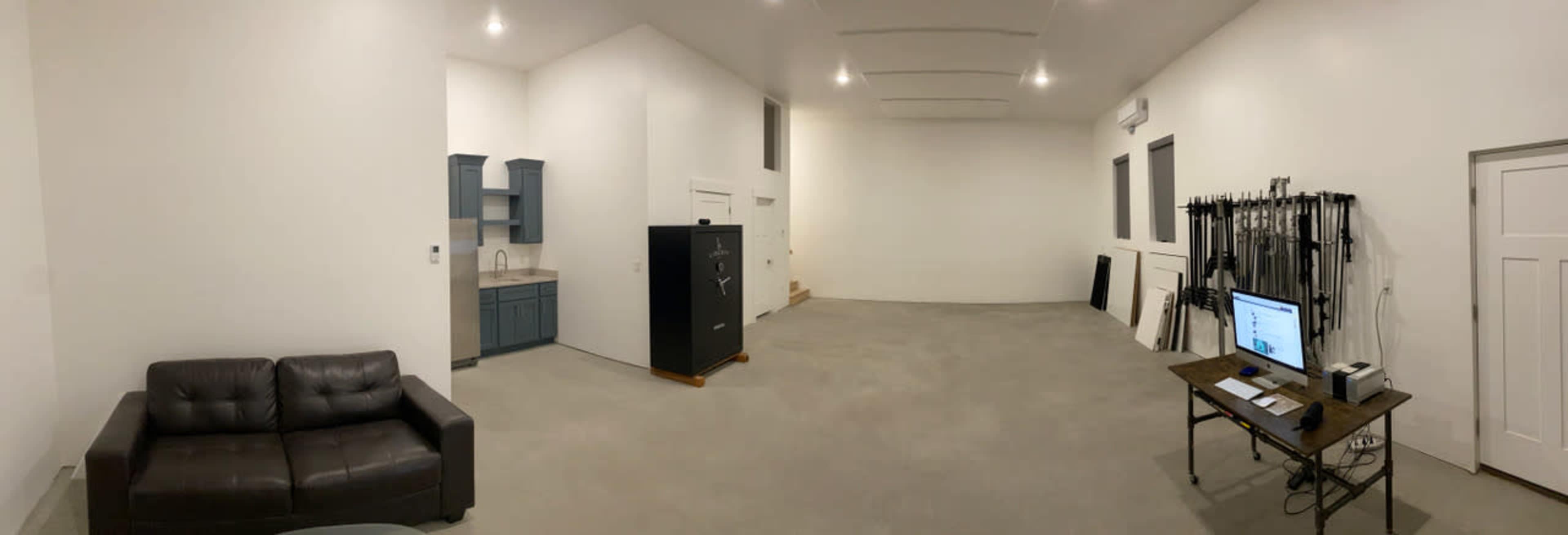 A spacious, empty room with a gray concrete floor, a black safe, a blue kitchenette, a brown couch, and a workstation with a computer on a wooden table.