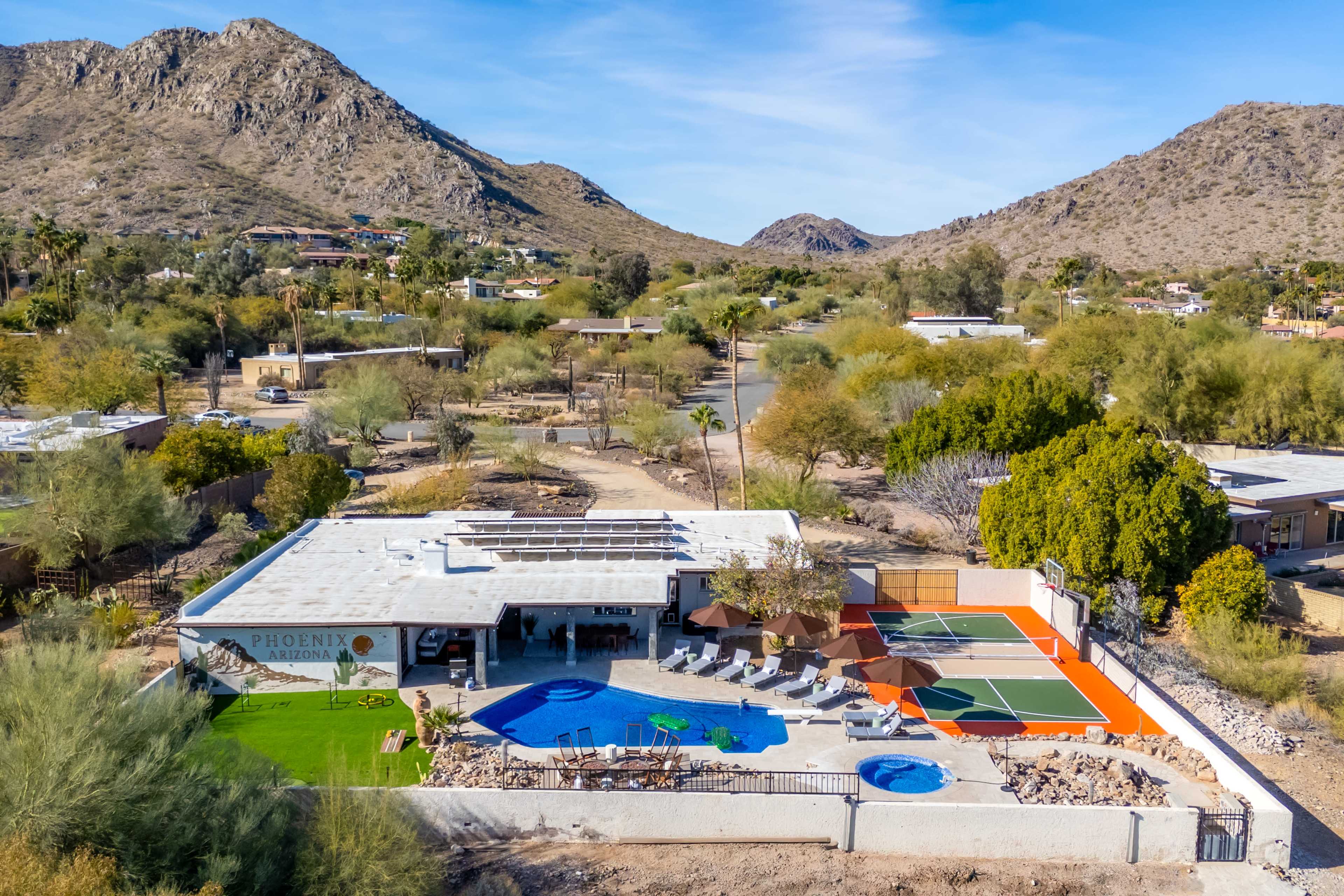 The image shows a modern house with a pool and two tennis courts, surrounded by desert landscape and mountains in the background.