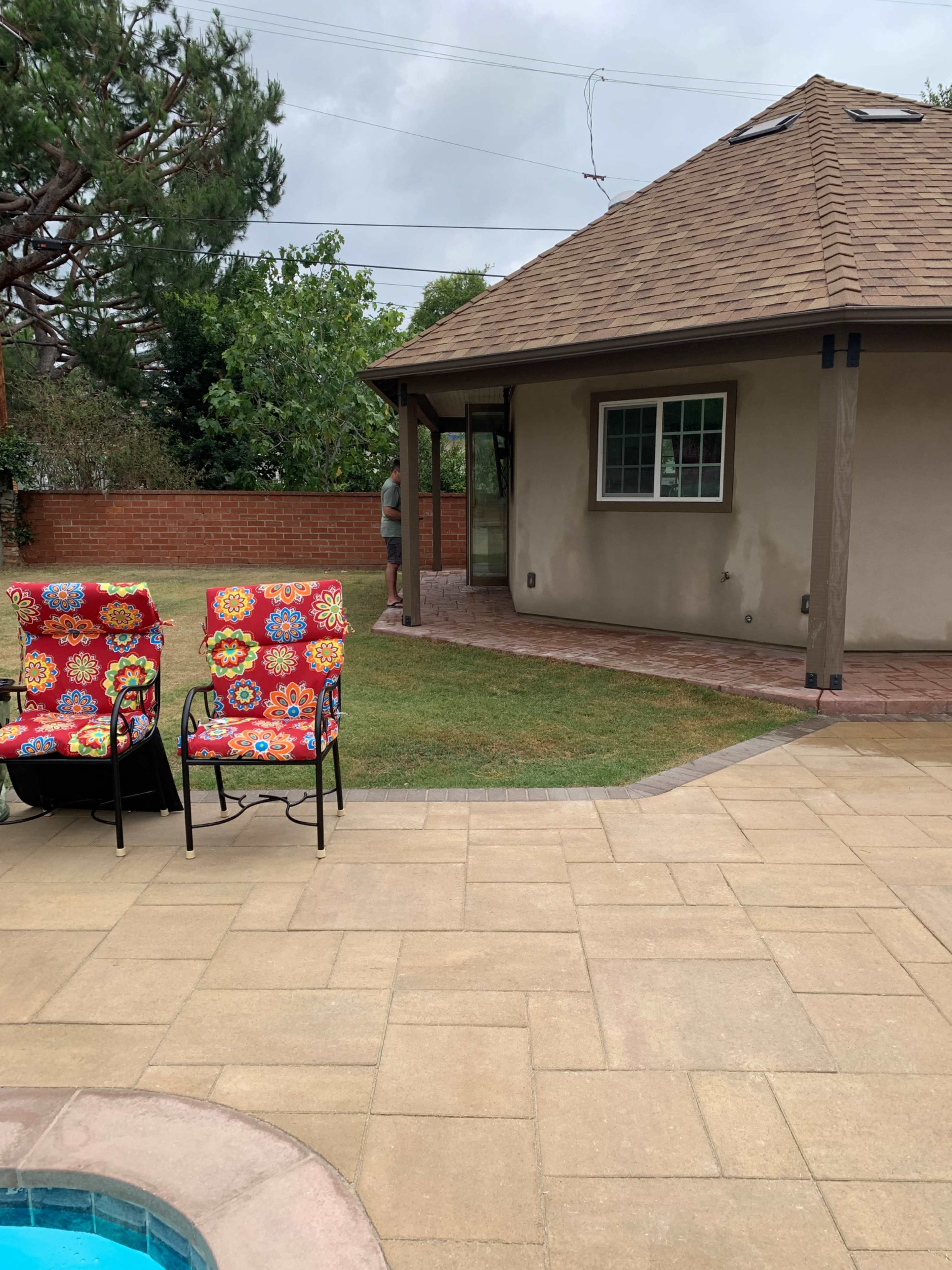 A backyard area with two patterned chairs facing a garden space, a brick walkway, and a house in the background.