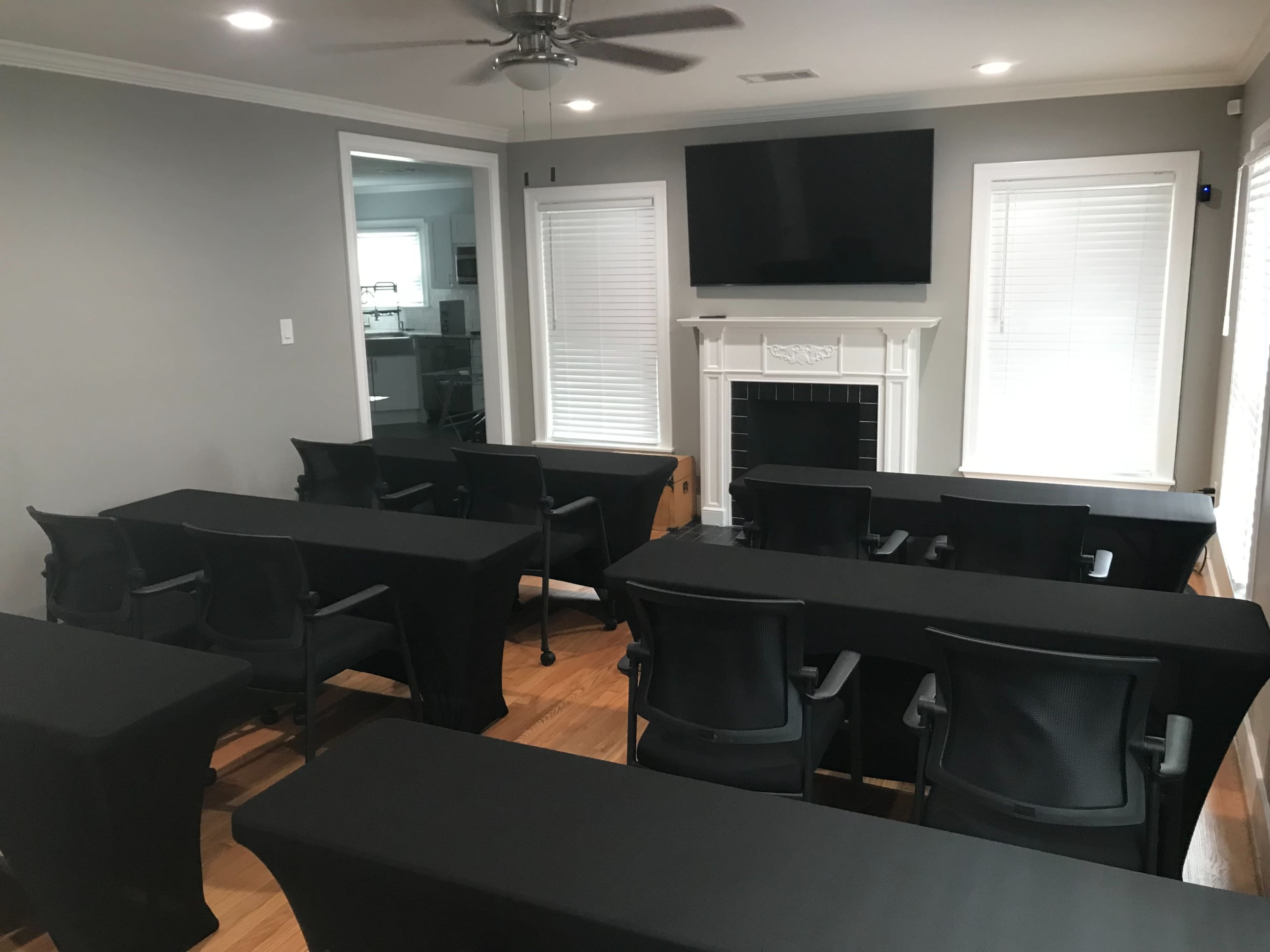 The image shows a conference room set up with several rows of tables covered in black cloth and chairs facing a wall-mounted TV above a fireplace.