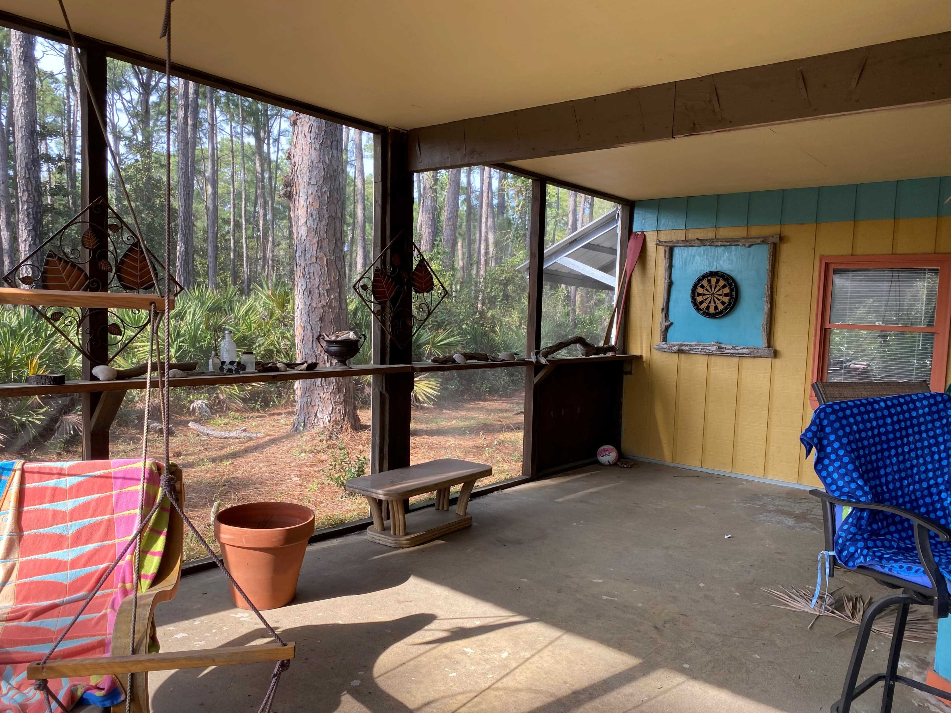 A screened-in porch with a dartboard mounted on the wall, a wooden bench, and a colorful chair, surrounded by trees in the background.